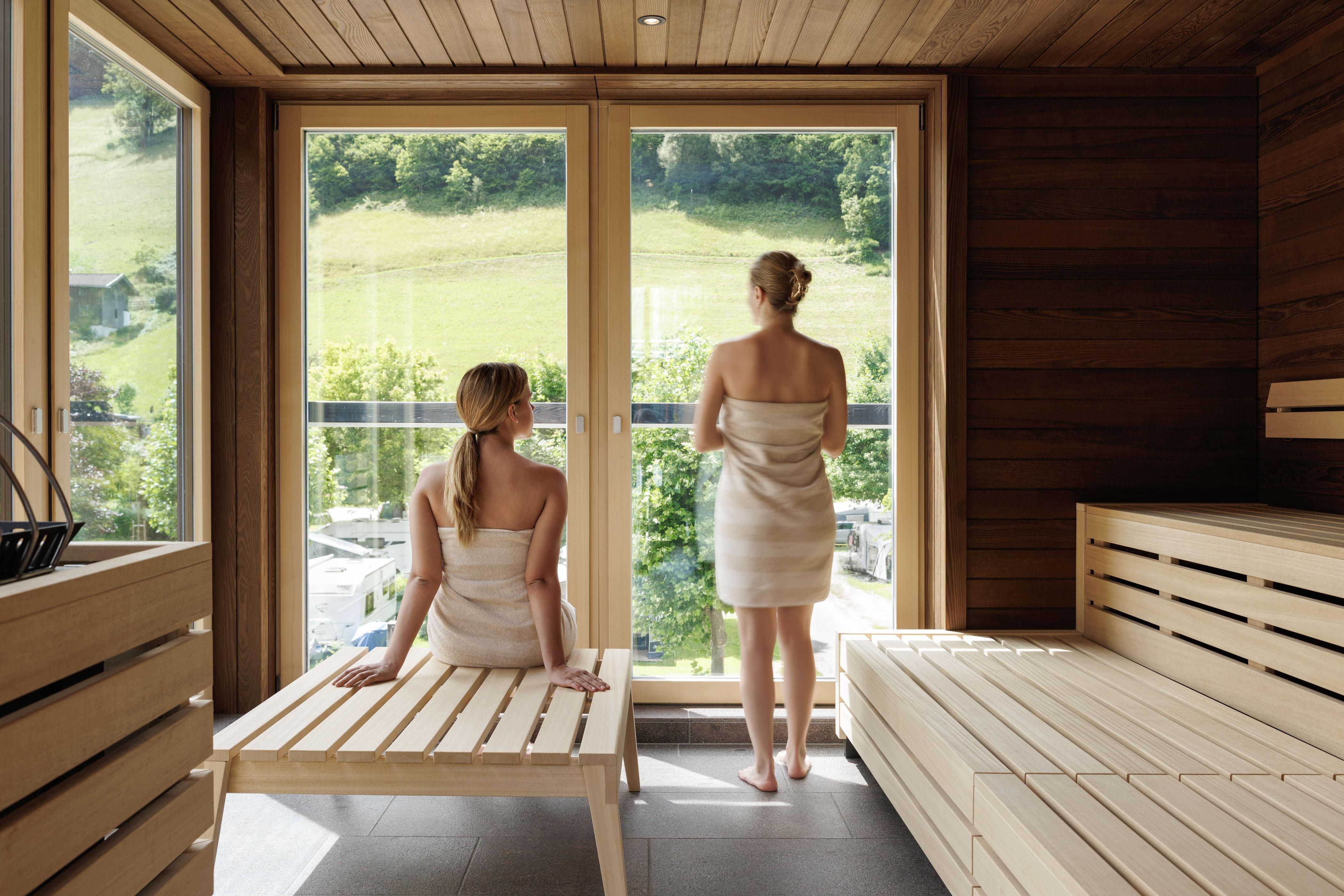 Zwei Frauen in einer Sauna schauen aus großen Fenstern auf die grüne Landschaft. Die Sauna ist aus Holz und hat eine entspannende Atmosphäre.