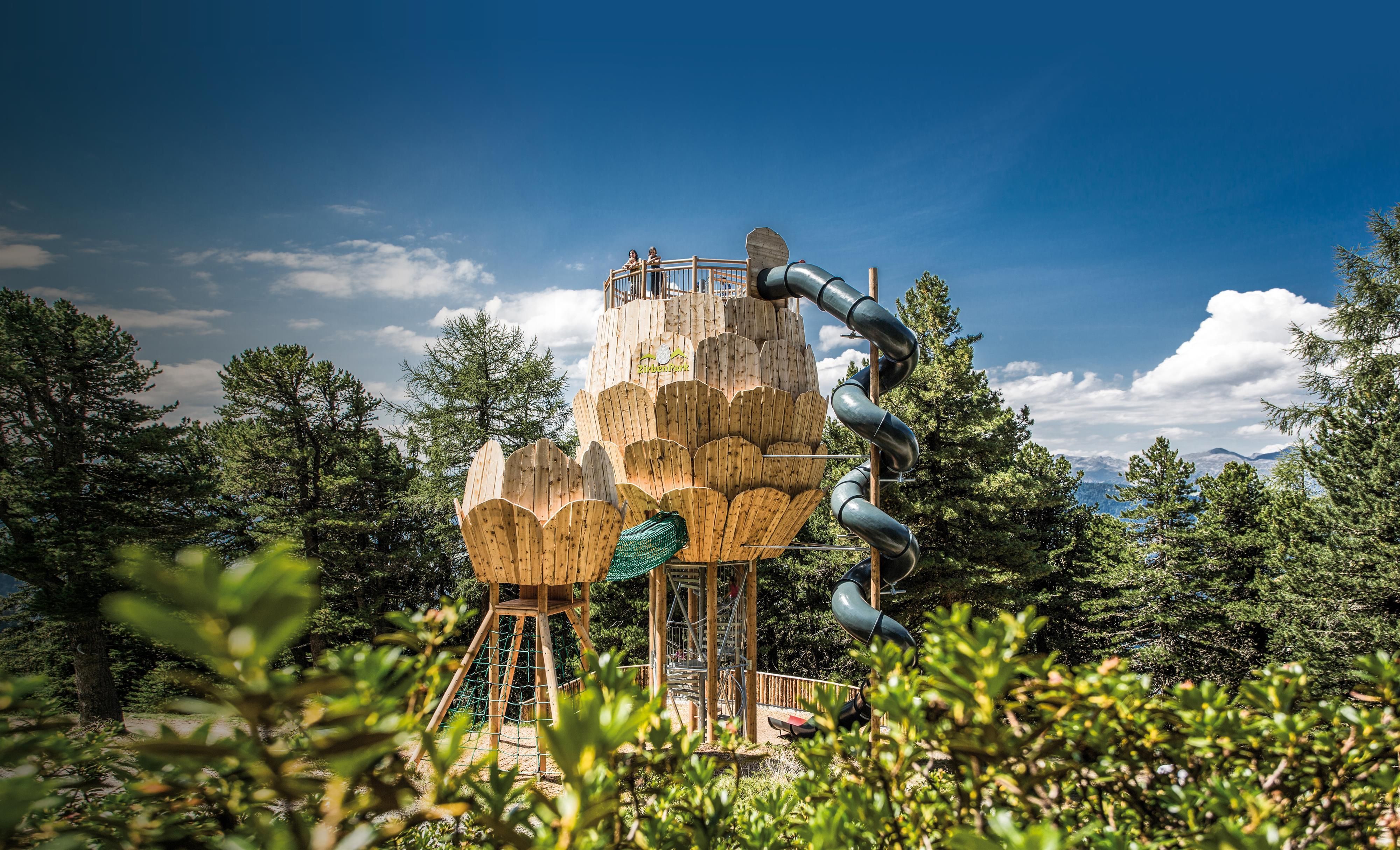 Ein kreativer Spielplatz in Baumhaus-Form mit einer Rutsche. Umgeben von grünen Bäumen und einem klaren Himmel.