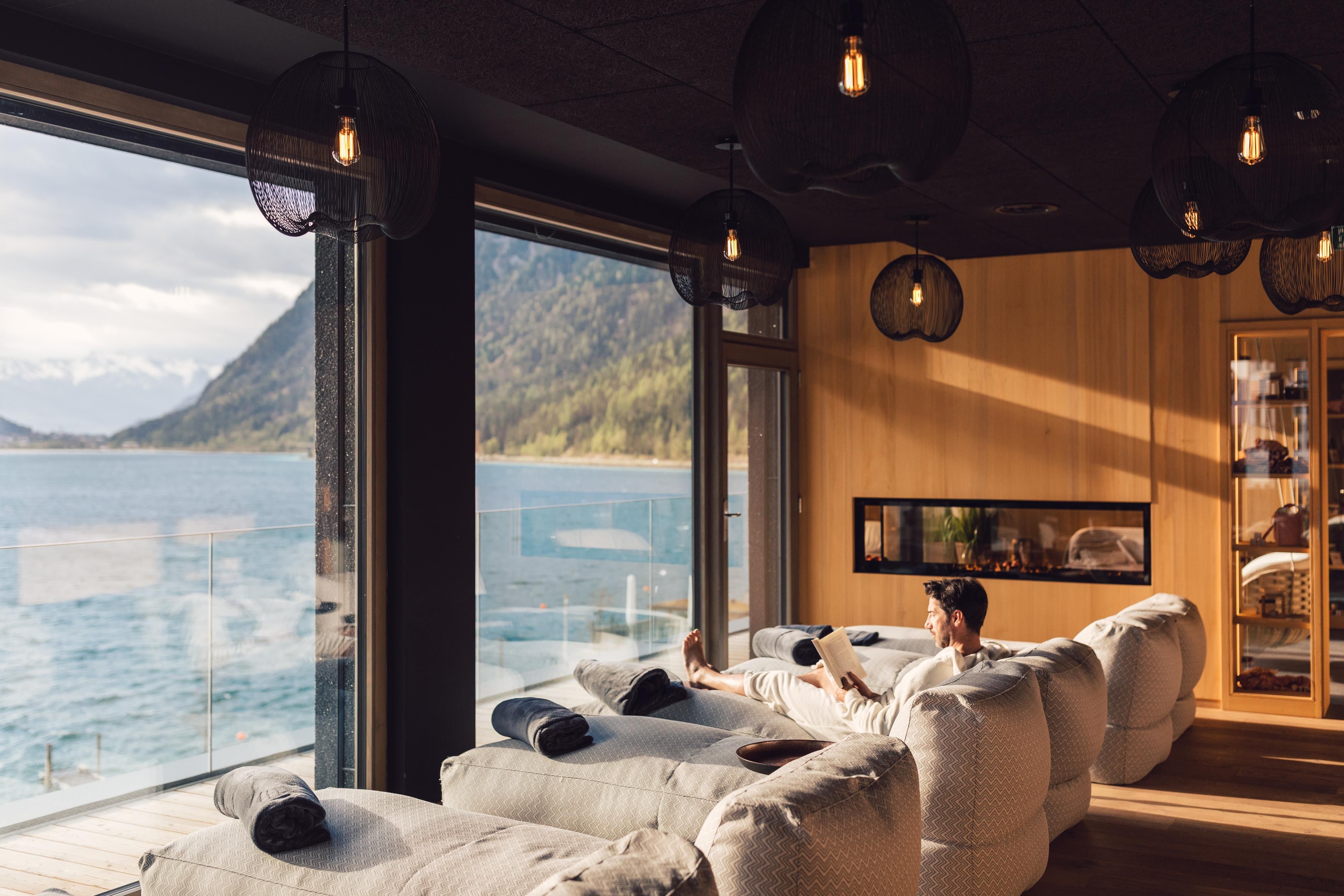Ein gemütlicher Raum mit großen Fenstern und Blick auf einen See. Eine Person entspannt auf einer bequemen Liege.