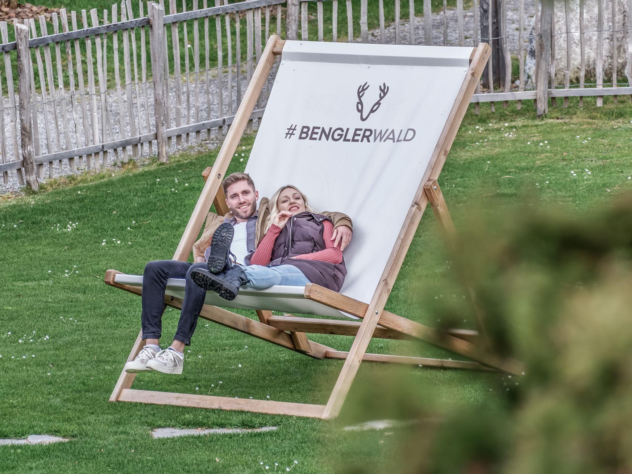 Ein großes Liegestuhl mit dem Schriftzug "#BENGLERWALD" steht auf einer grünen Wiese. Zwei Personen entspannen darauf und genießen die Zeit im Freien.