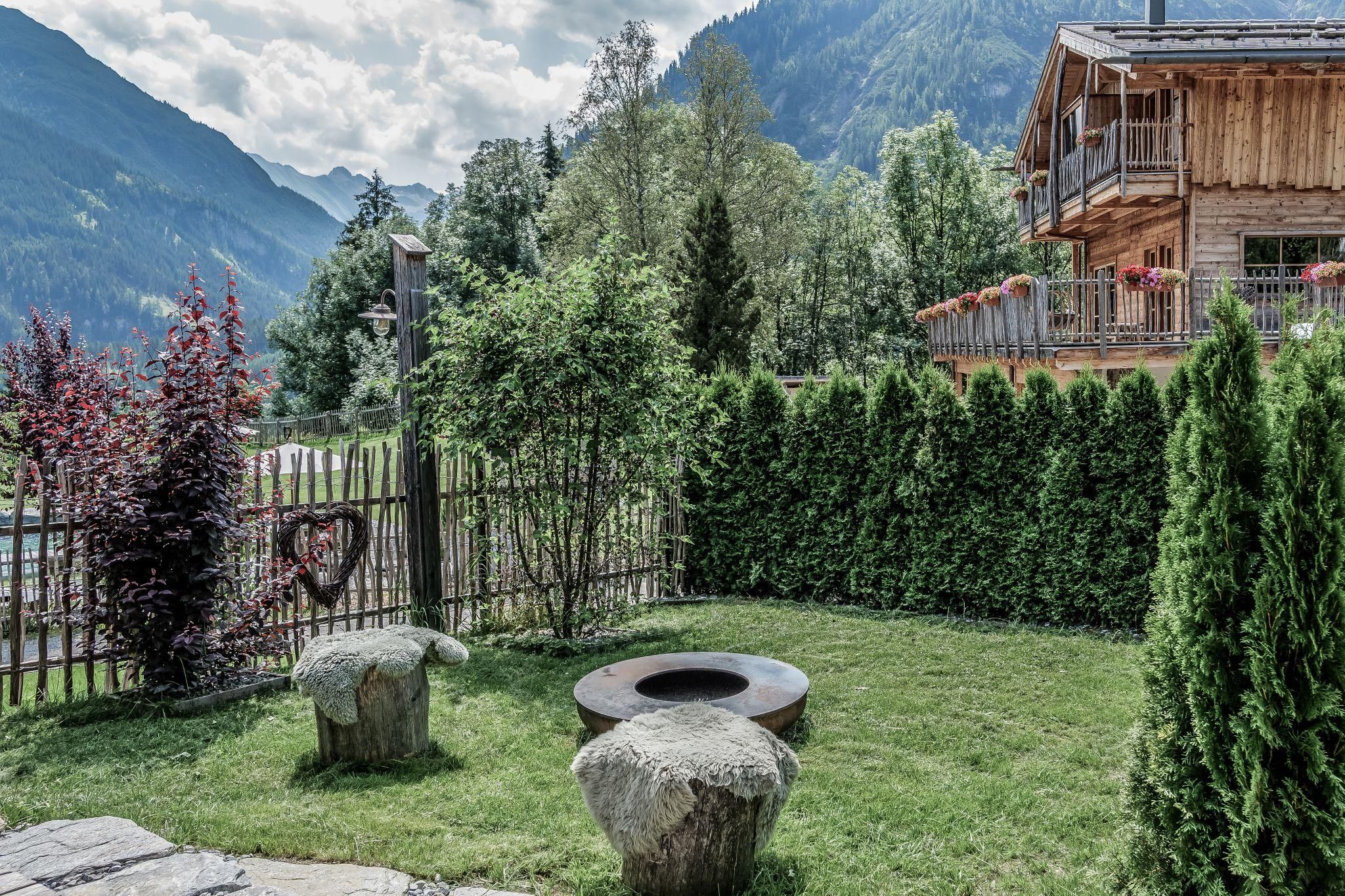 Ein schöner Garten mit grünen Pflanzen und einem kleinen Tisch aus Stein. Im Hintergrund ist ein Chalet und Berge zu sehen.