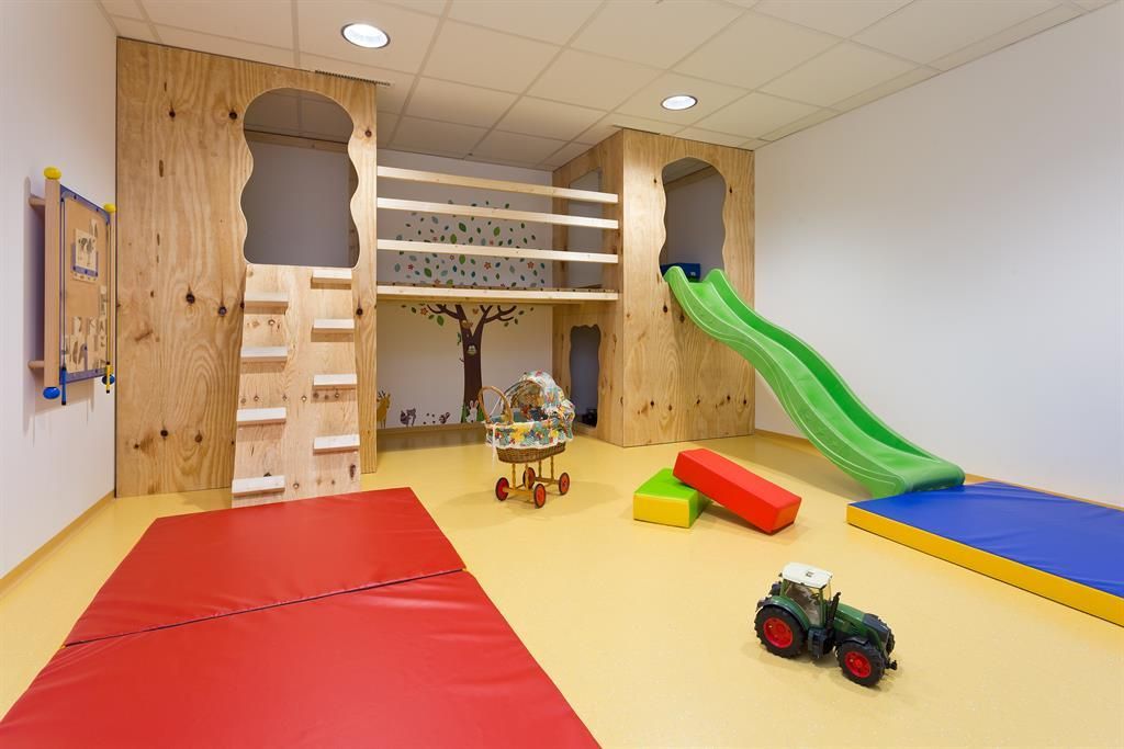 Ein farbenfroher Spielraum mit einer Rutsche und Treppe aus Holz. Spielzeuge und Matten sorgen für eine einladende Umgebung zum Spielen.