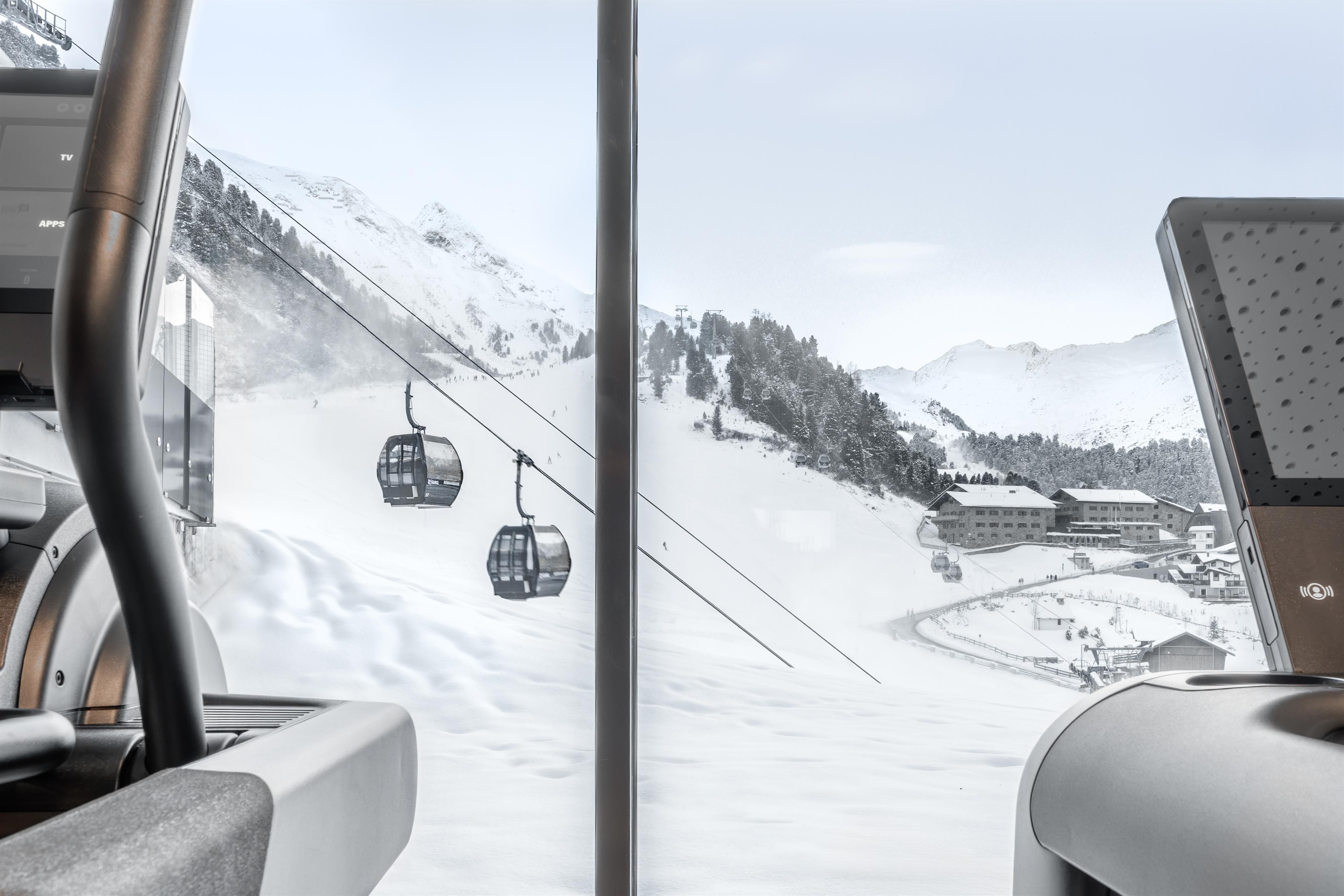 Eine Aussicht auf schneebedeckte Berge mit einer Gondelbahn im Hintergrund. Im Vordergrund sind moderne Fitnessgeräte sichtbar.