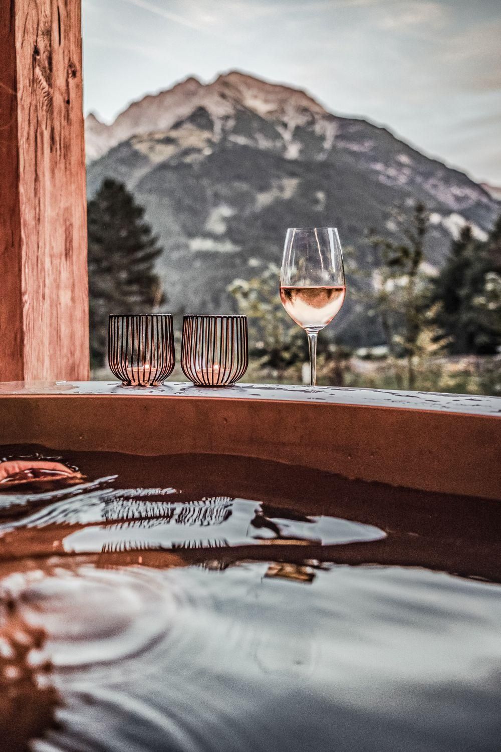 Ein entspannendes Bad im Hot Pot mit einer Aussicht auf die Berge. Auf dem Rand stehen Weingläser und eine ruhige Atmosphäre ist spürbar.