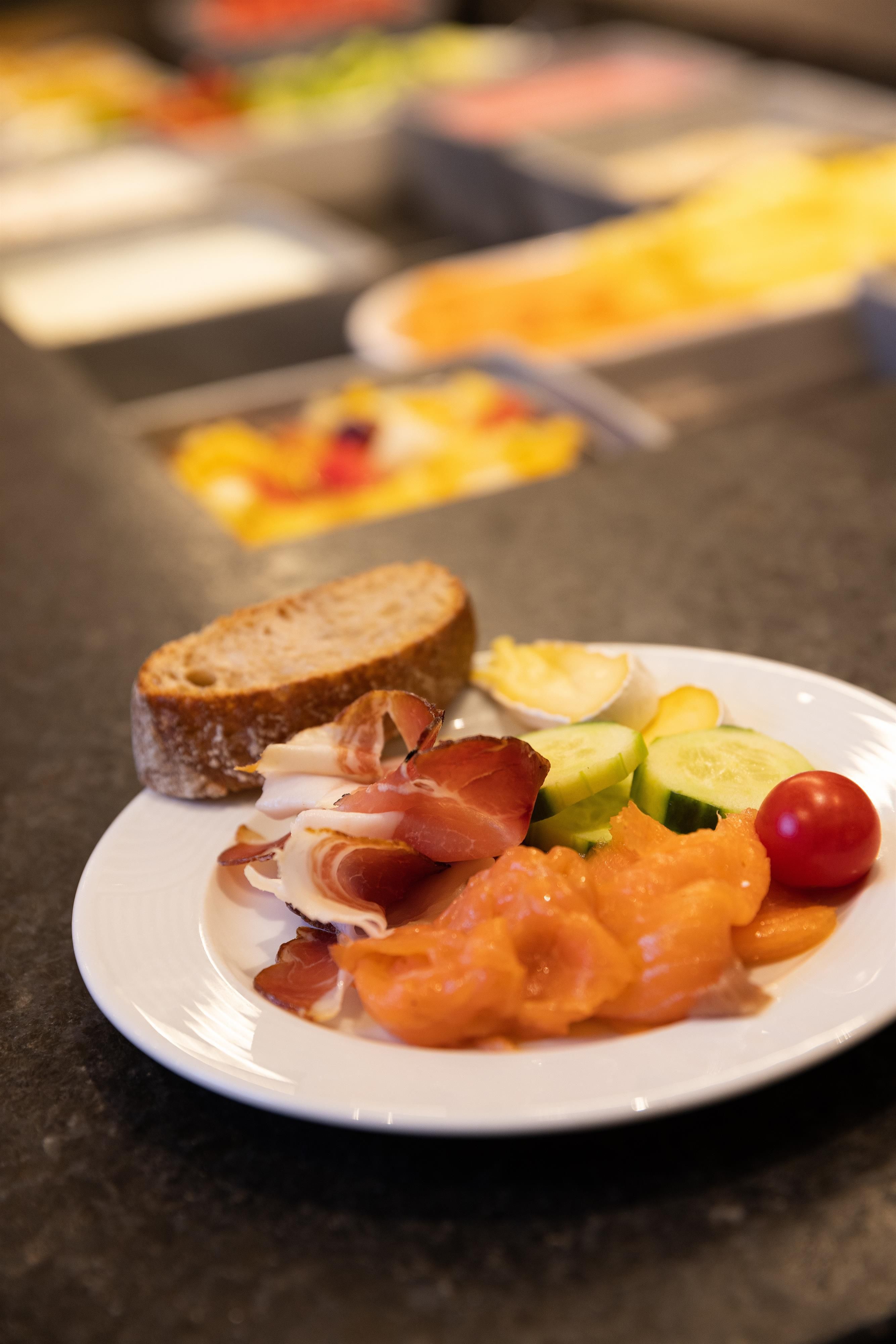 Ein Teller mit verschiedenen Lebensmitteln, darunter Brot, Aufschnitt, Lachs und Gemüse. Im Hintergrund sind weitere Speisen sichtbar.