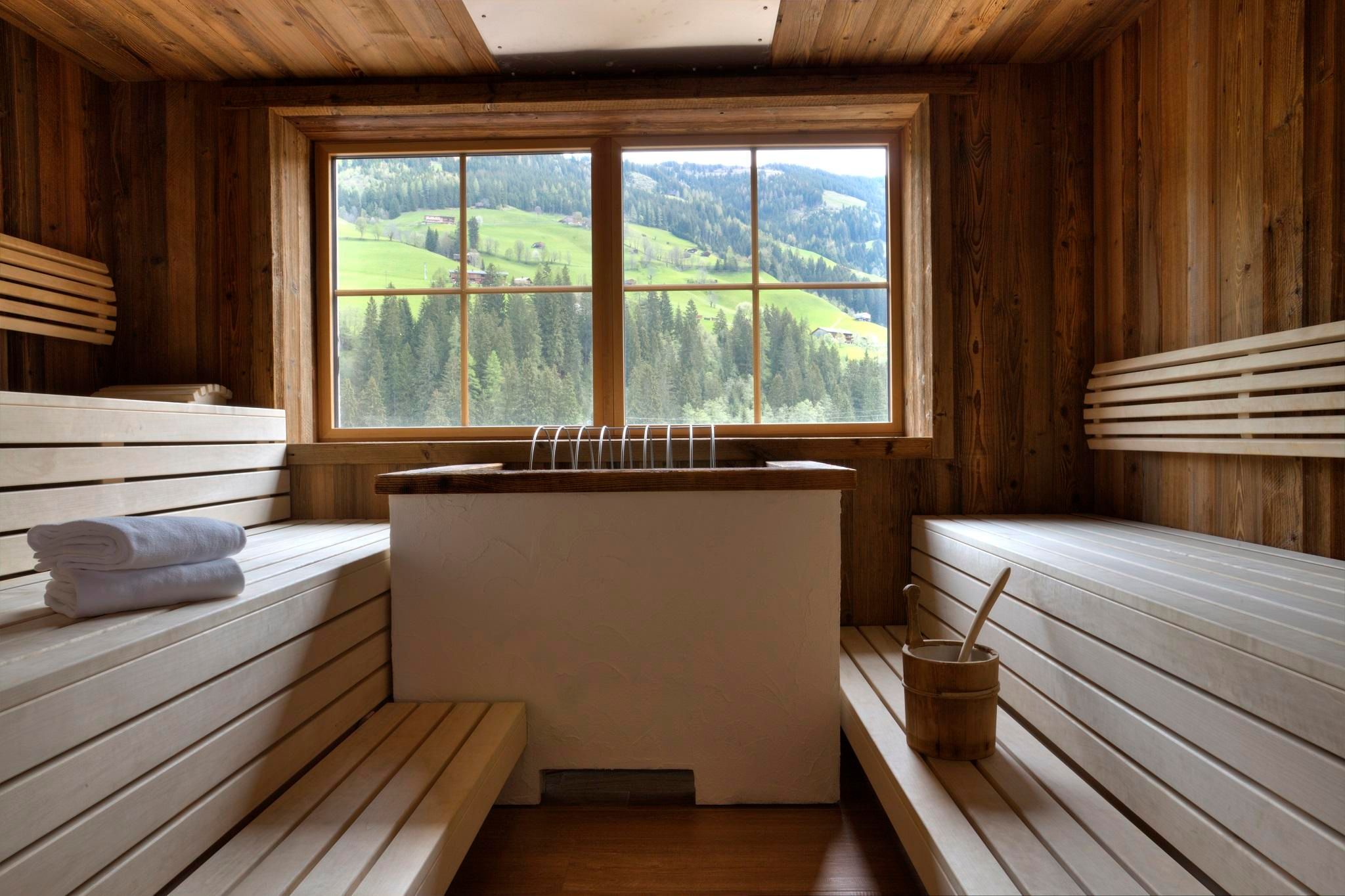 Ein heller Sauna-Raum mit Holzverkleidung und einer großen Fensterfront. Der Blick reicht über grüne Hügel und Wälder.
