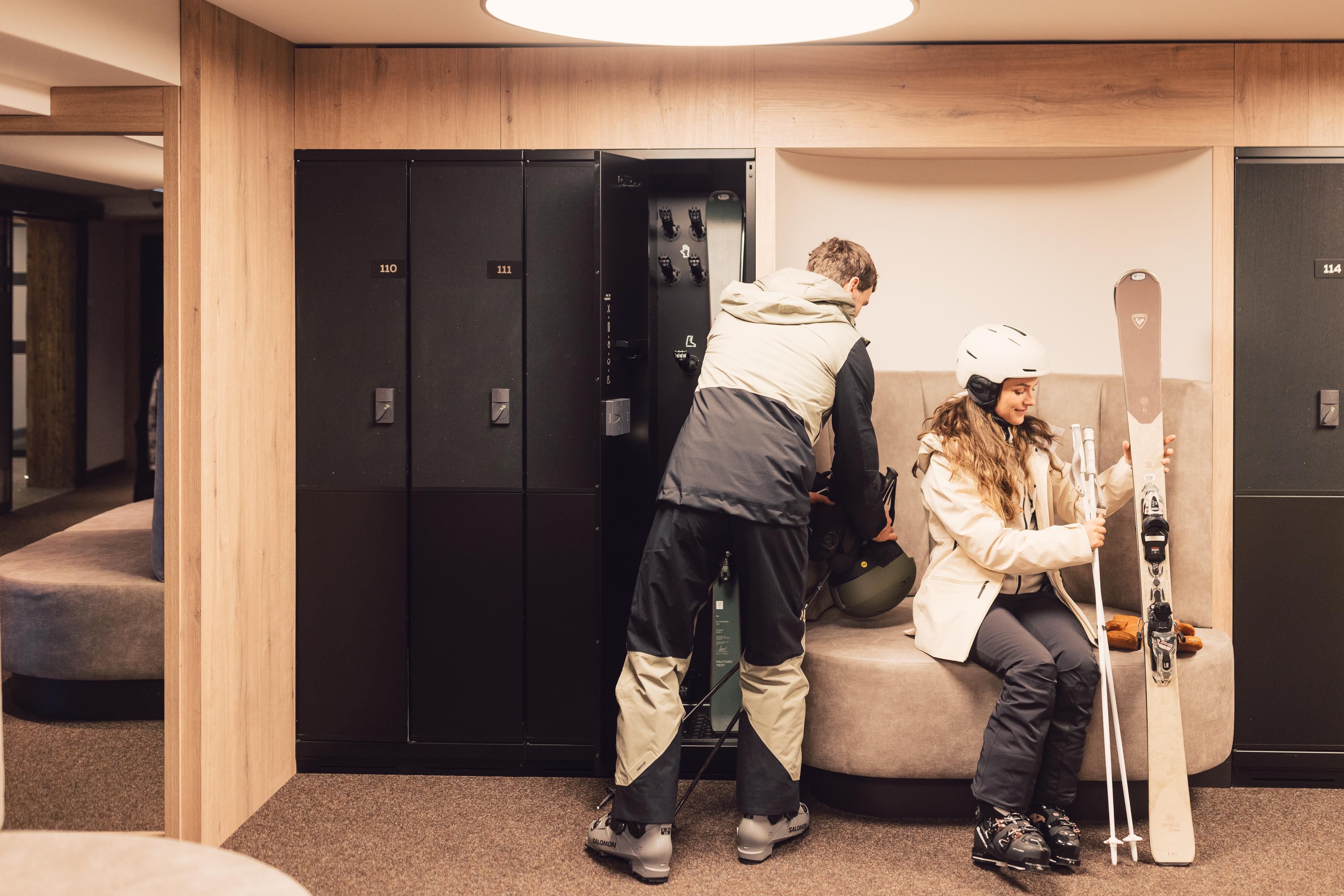 Eine gemütliche Skiausrüstungsumkleide mit zwei Personen. Eine Frau sitzt mit Skiern, während ein Mann an einem Spind steht.