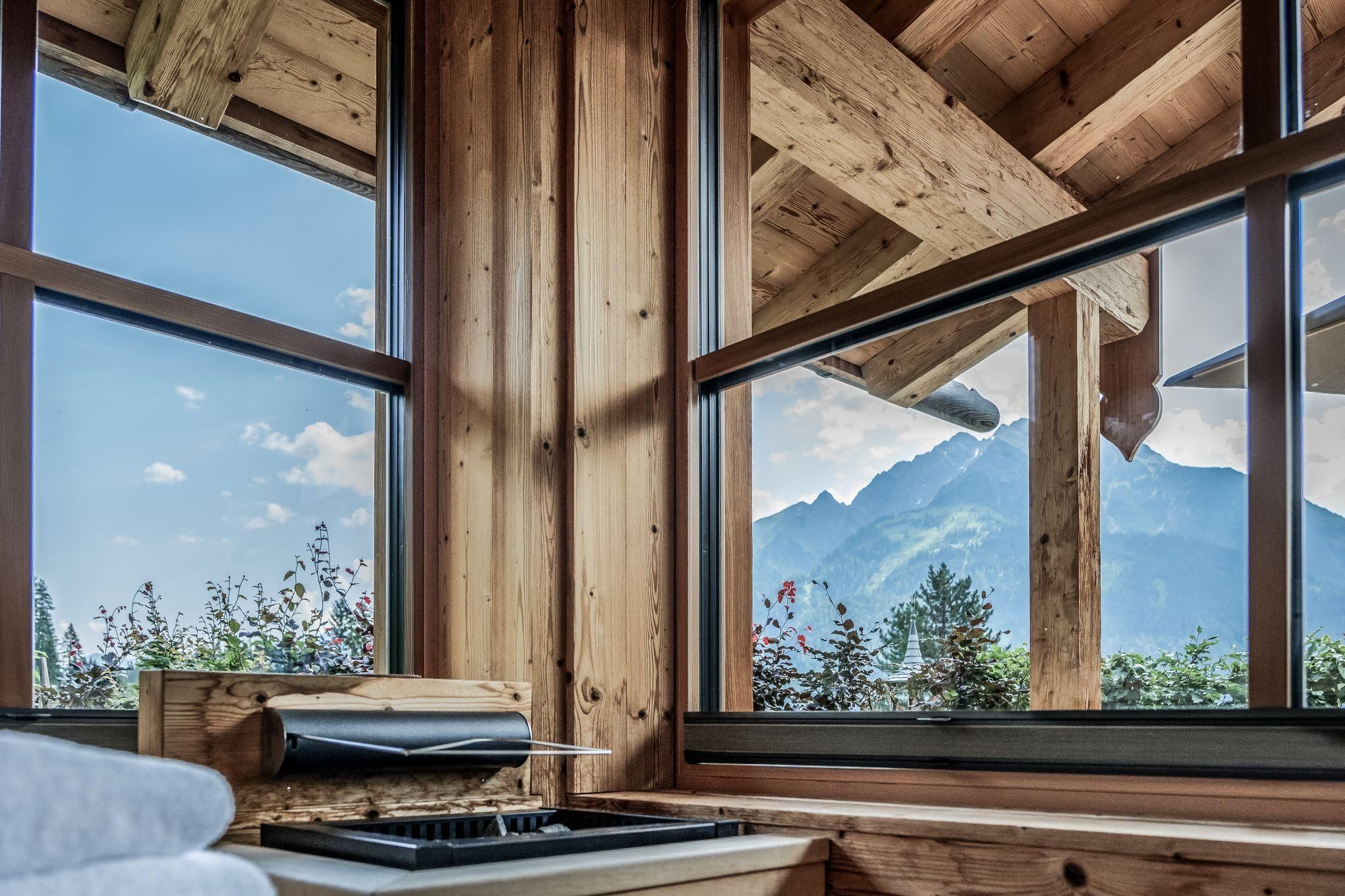Ein gemütlicher Innenraum mit Holzwänden und großen Fenstern. Im Hintergrund sind majestätische Berge und ein blauer Himmel zu sehen.
