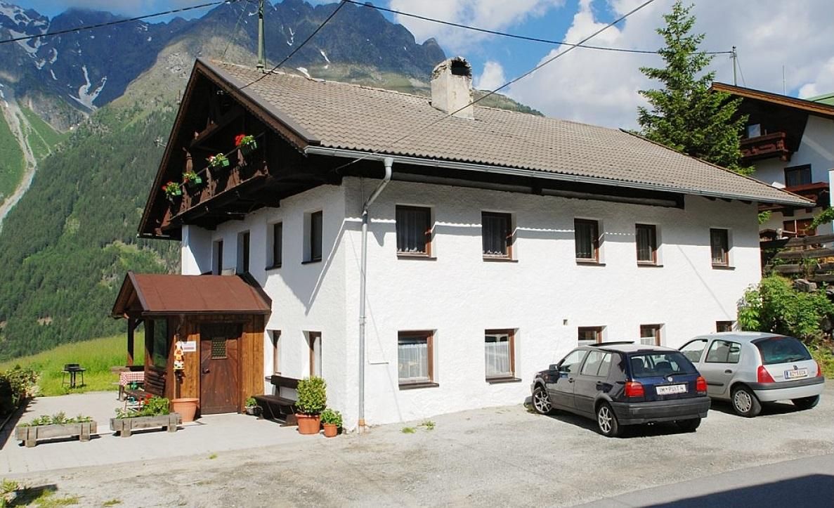 Ein gemütliches, zwei-geschossiges Haus im alpinen Stil, umgeben von Bergen. Vor dem Haus stehen mehrere Autos und es gibt Blumenkästen an den Fenstern.