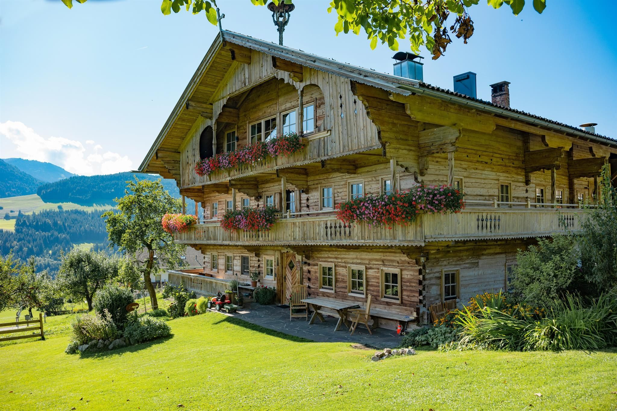 Ein charmantes Holzhaus mit mehreren Balkonen und bunten Blumenkästen. Es umgeben eine grüne Wiese und malerische Berge im Hintergrund.