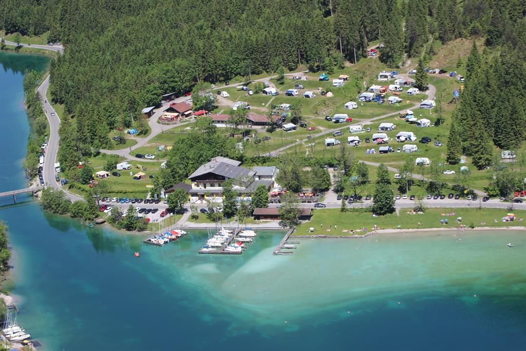 Ein malerischer Campingplatz am Wasser mit grünen Wiesen und vielen Wohnwagen. Im Vordergrund ist ein schöner See mit Booten und klaren Wasser zu sehen.