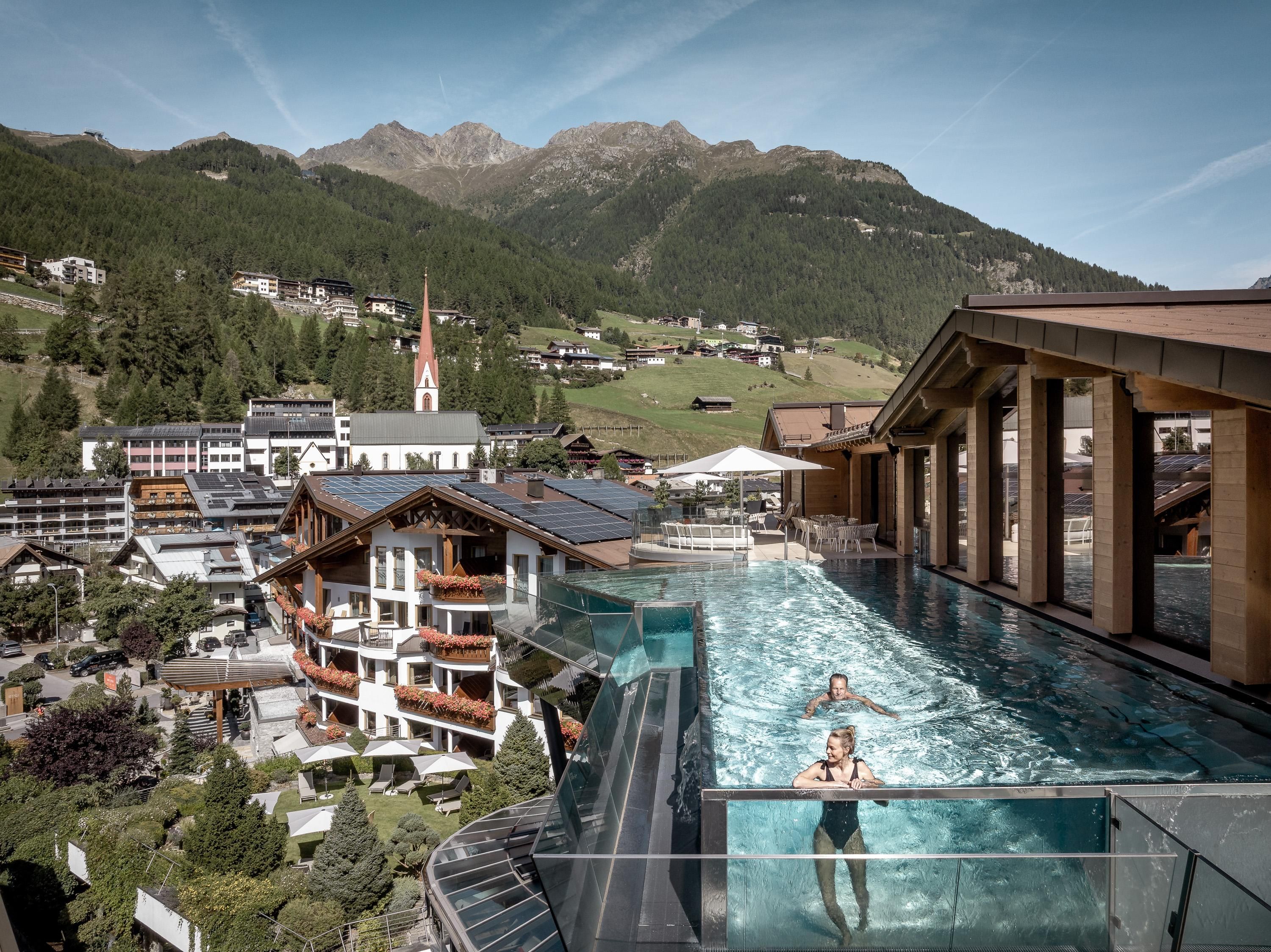 Ein Infinity-Pool mit Blick auf die Berge und die umliegenden Gebäude. Im Wasser schwimmen zwei Personen, während die Sonne scheint.
