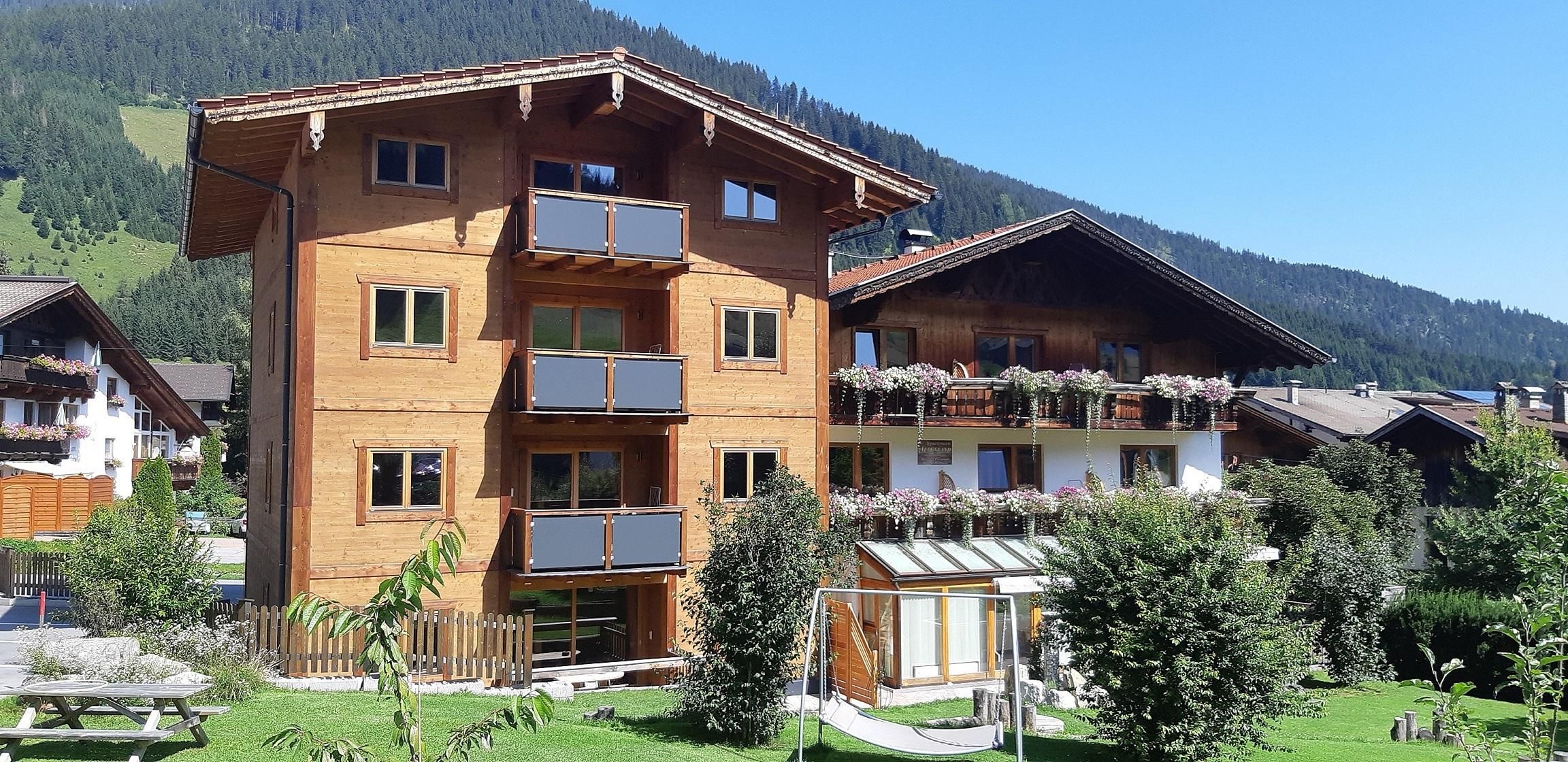 Ein traditionelles Holzhaus mit mehreren Balkonen und schöner Blumendekoration. Umgeben von grüner Landschaft und Bäumen, vermittelt es eine einladende Atmosphäre.