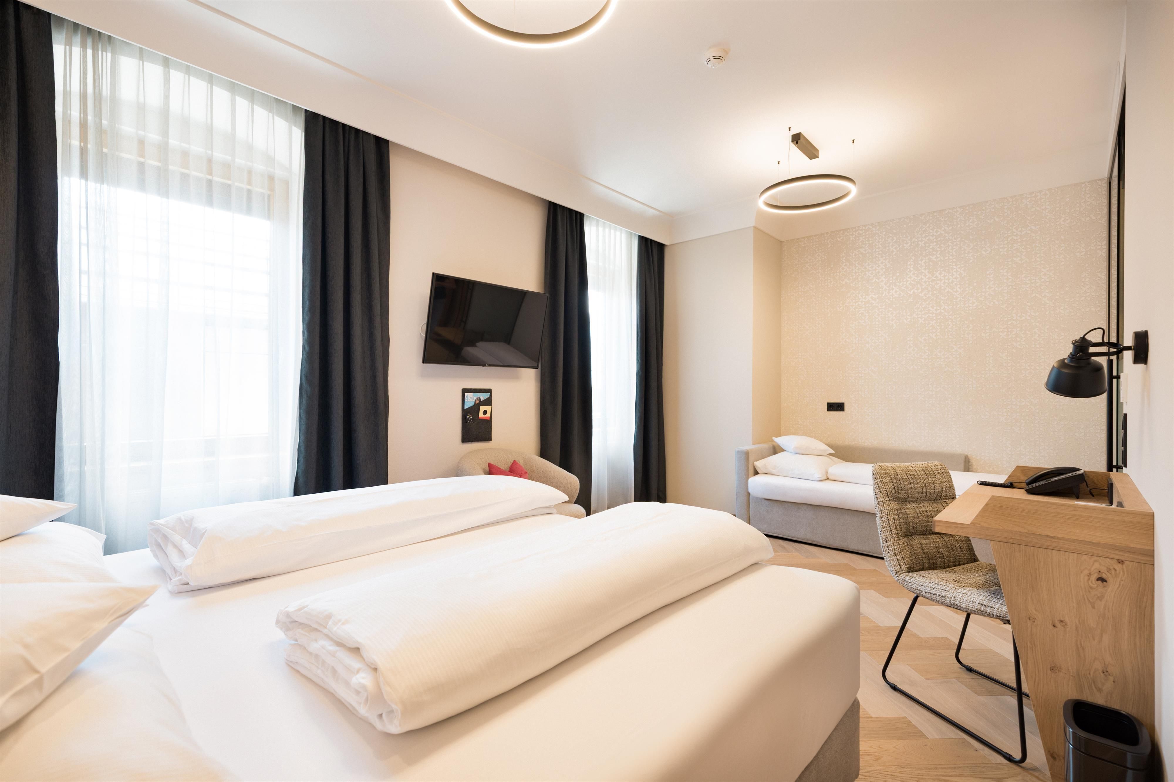 Ein modernes Hotelzimmer mit zwei Betten, einem Schreibtisch und einem Fernseher. Helle Vorhänge sorgen für eine freundliche Atmosphäre.