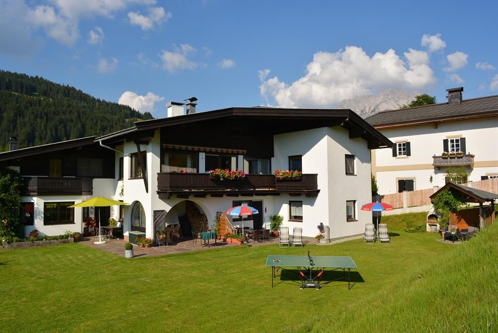 Ein gemütliches Haus mit Balkon und einer großflächigen Wiese. Auf dem Grundstück stehen Tische mit Sonnenschirmen und eine Tischtennisplatte.