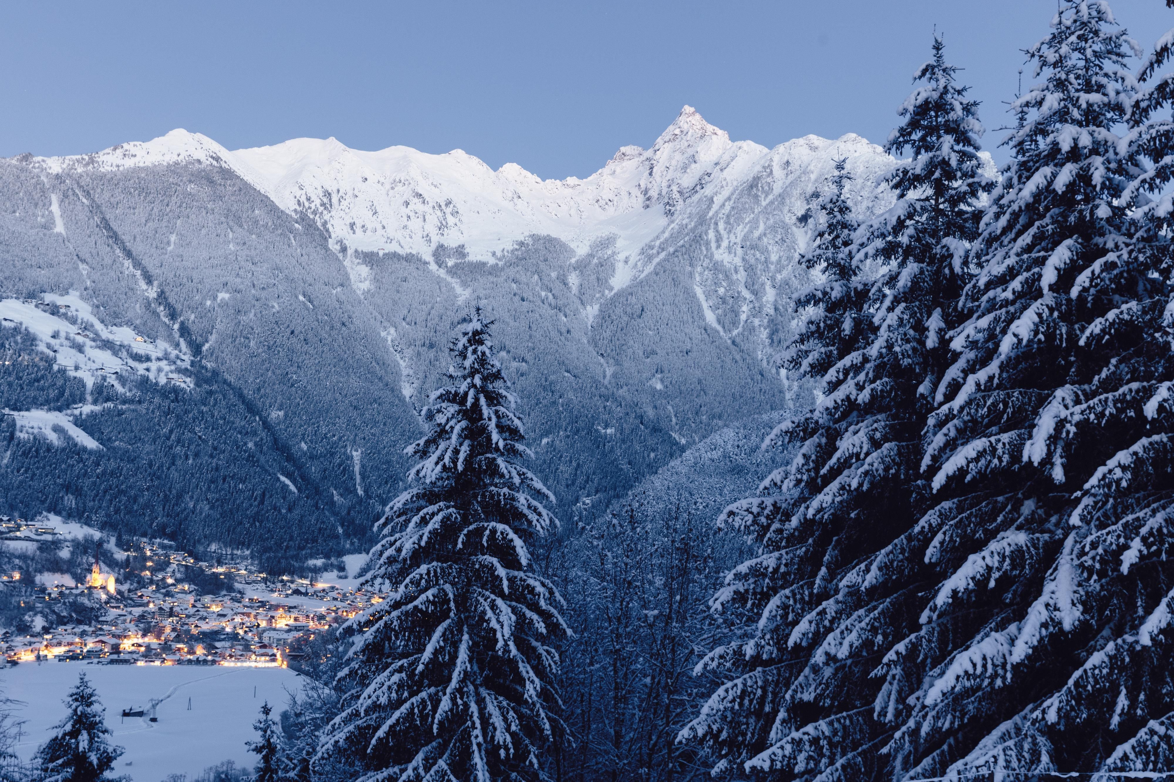 Eine beeindruckende winterliche Berglandschaft mit schneebedeckten Gipfeln. Im Vordergrund stehen hohe Tannenbäume, während das Licht der Stadt in der Talsohle schimmert.