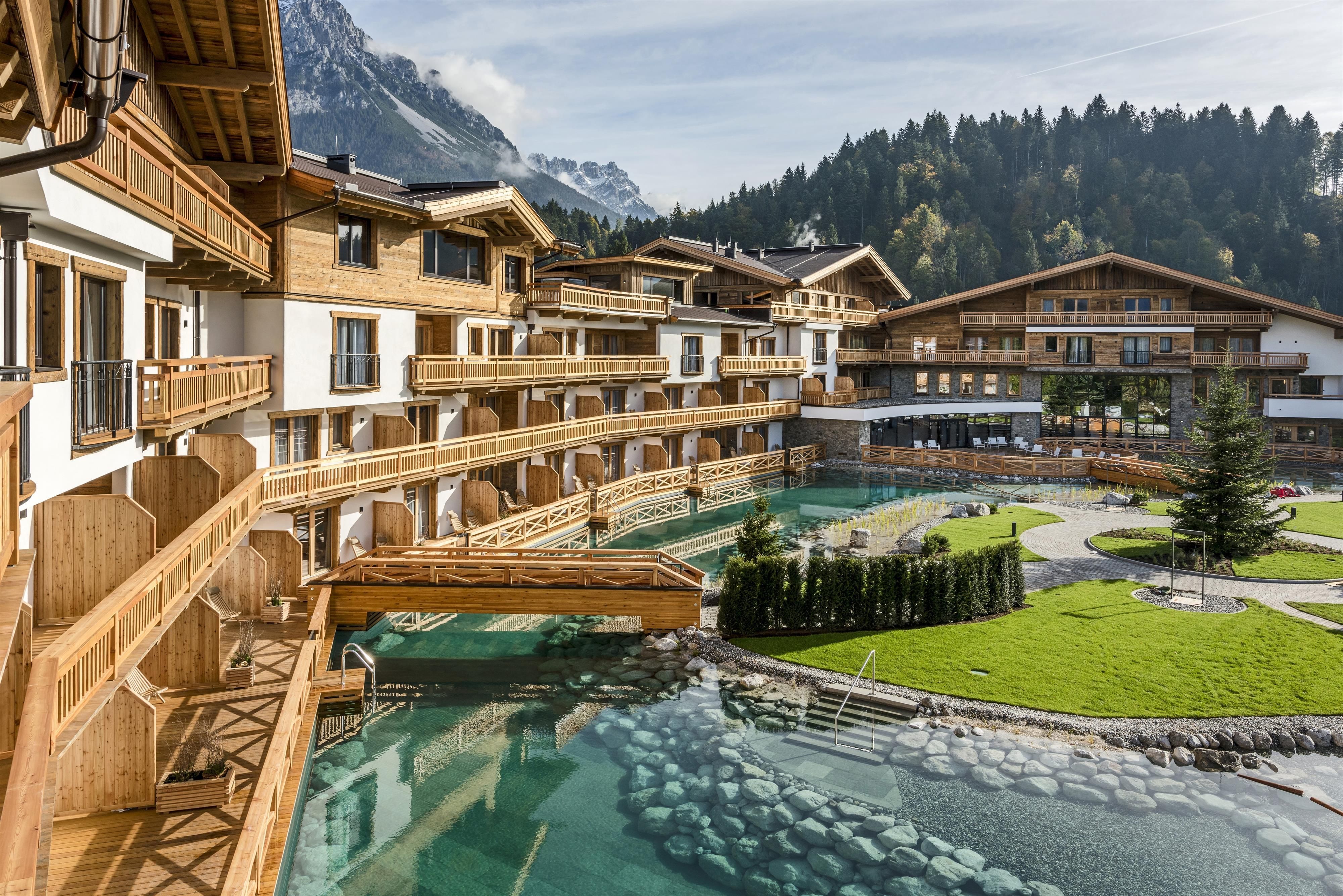 Ein schönes Wellness-Resort mit Holzarchitektur und einem klaren Wasserlauf. Im Hintergrund sind die Berge und ein grüner Bereich mit Rasen und Pflanzen zu sehen.