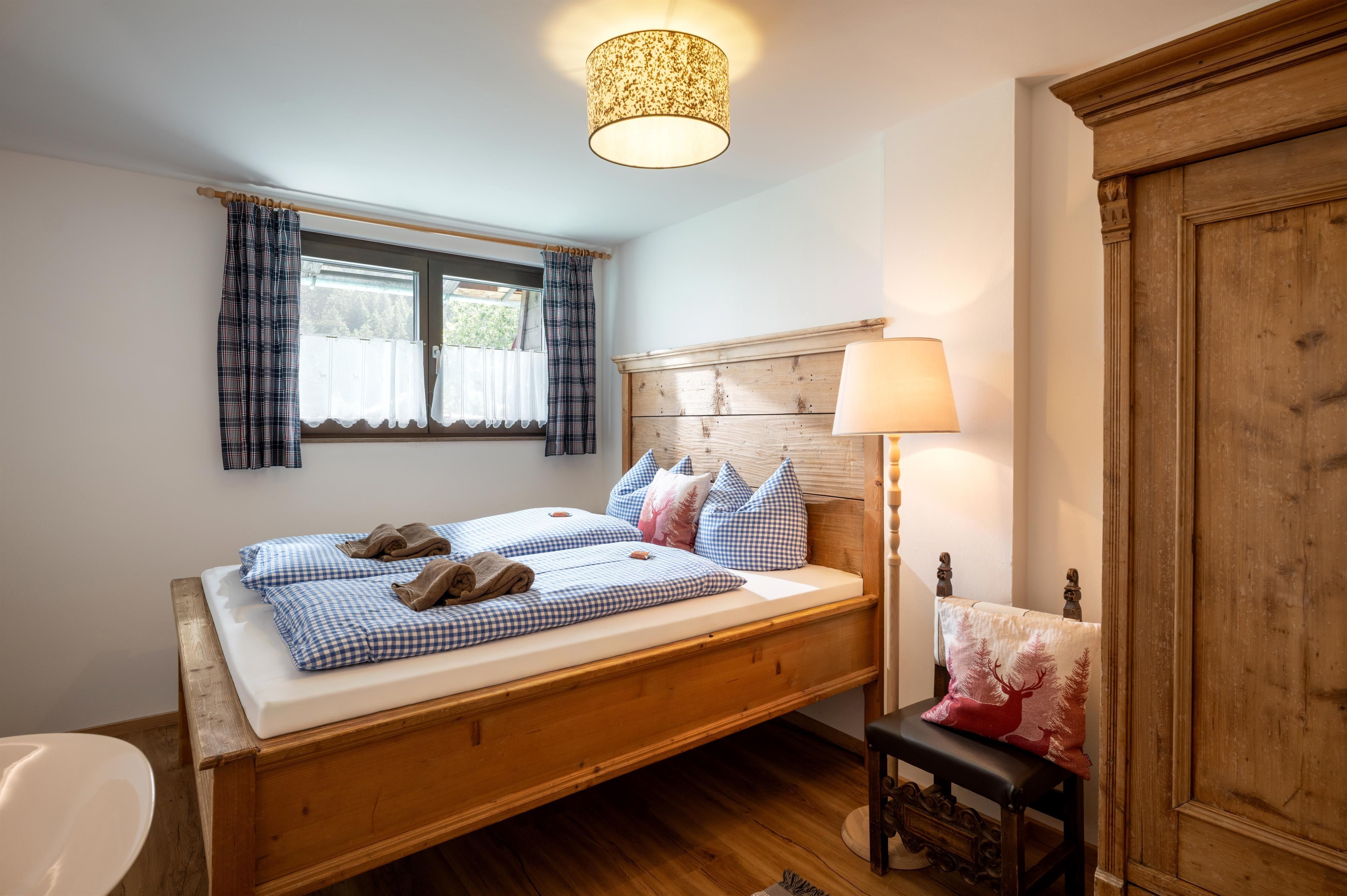 Ein gemütliches Schlafzimmer mit einem Holzbett und blauen Kissen. Der Raum hat ein Fenster mit Vorhängen und eine warmen Beleuchtung.