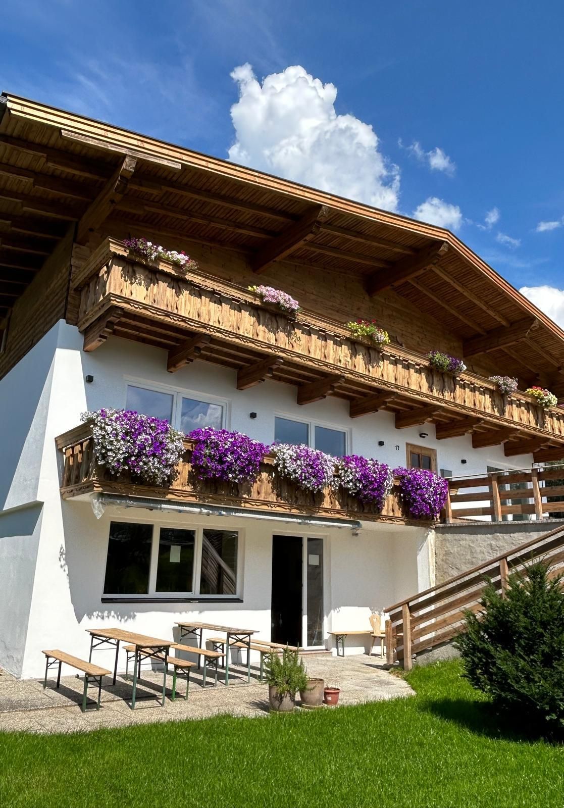 Ein charmantes Haus mit Blumen auf den Balkonen. Der Himmel ist blau und es gibt viel Grünfläche rundum.