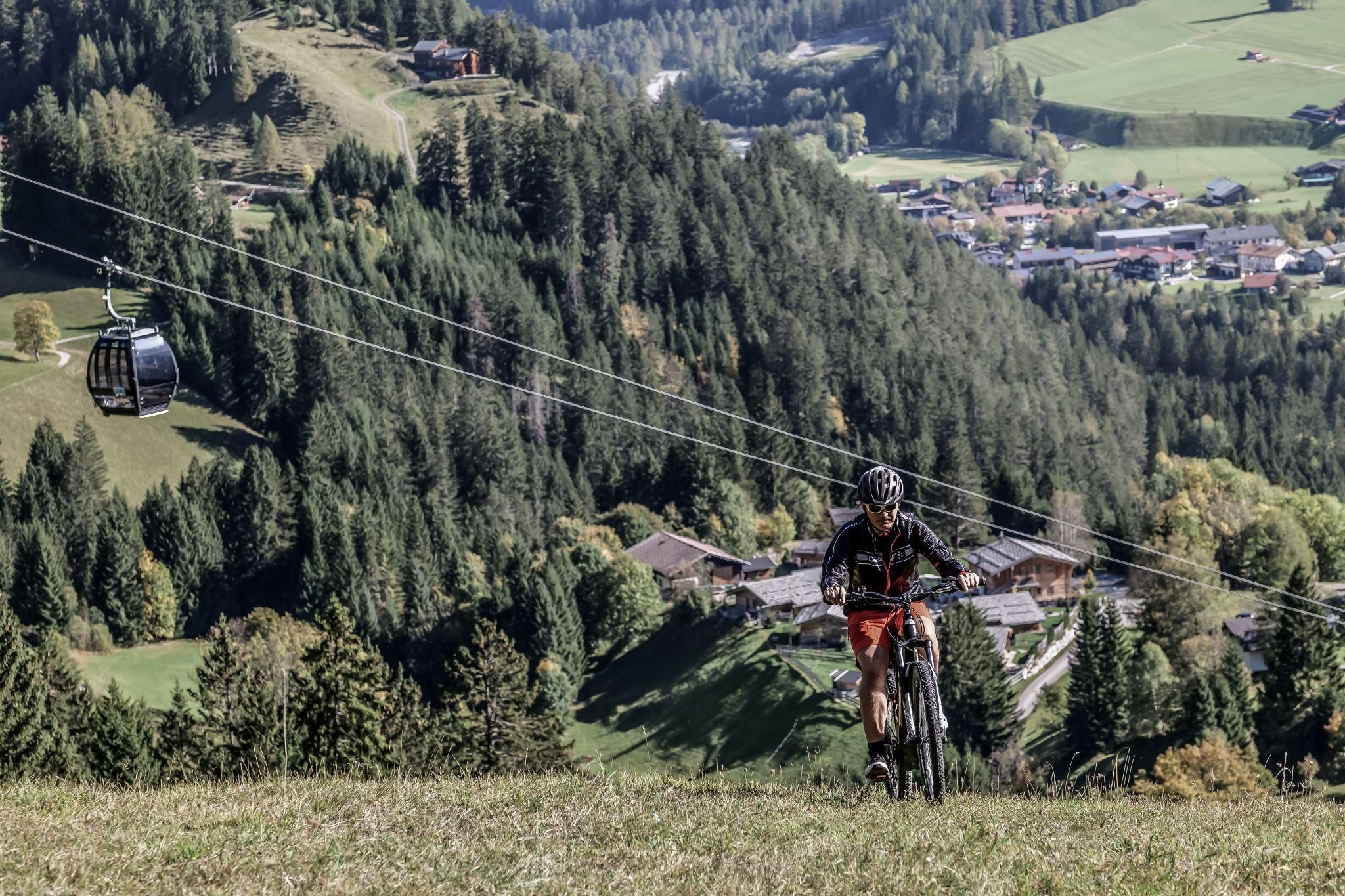 Ein Radfahrer fährt auf einem grünen Hügel in den Bergen. Im Hintergrund sind eine Seilbahn und ein malerisches Chalet Dorf zu sehen.