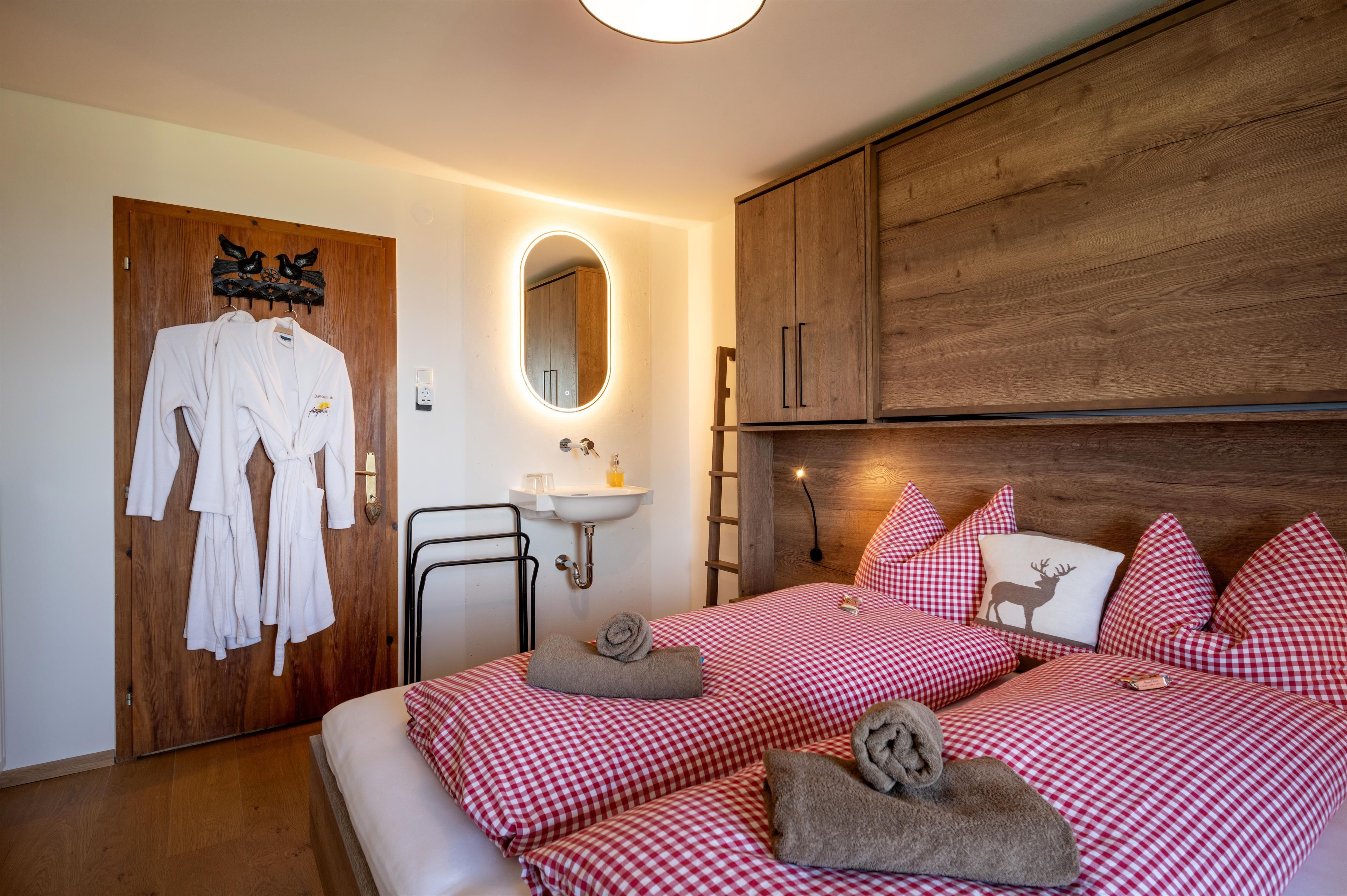 Ein gemütliches Schlafzimmer mit zwei Einzelbetten und rot-weißem Karomuster. Es gibt einen Spiegel, einen kleinen Waschbereich und Bademäntel hängen an der Tür.