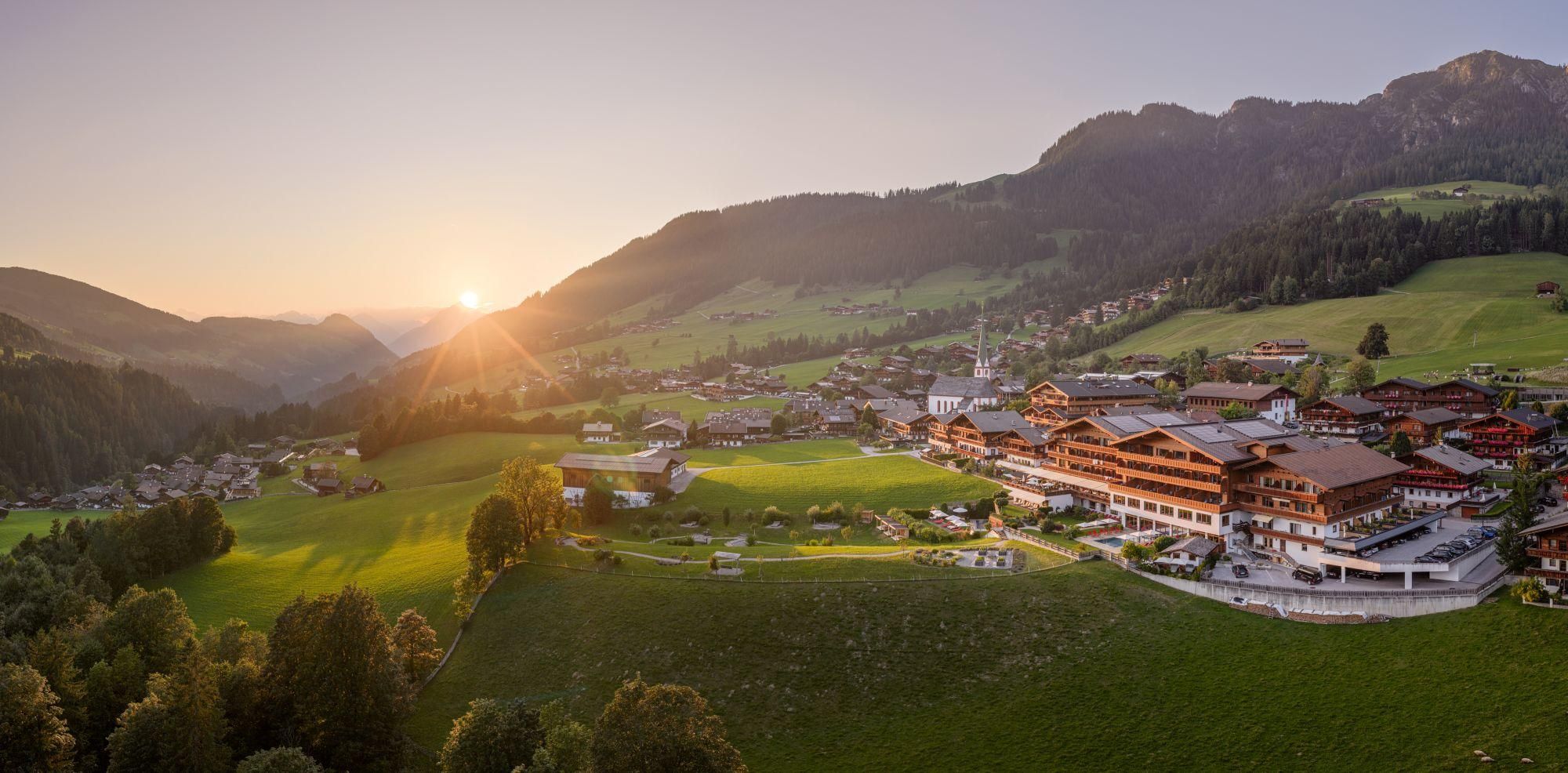 Eine malerische Berglandschaft mit sanften Hügeln und einem kleinen Dorf. Die Sonne geht über den Bergen auf und taucht die Szene in warmes Licht.