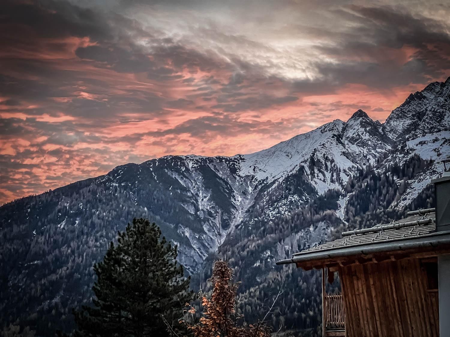 Die Aussicht zeigt schneebedeckte Berge unter einem bunten Himmel bei Sonnenuntergang. Im Vordergrund ist ein Chalet und einige Bäume sichtbar.