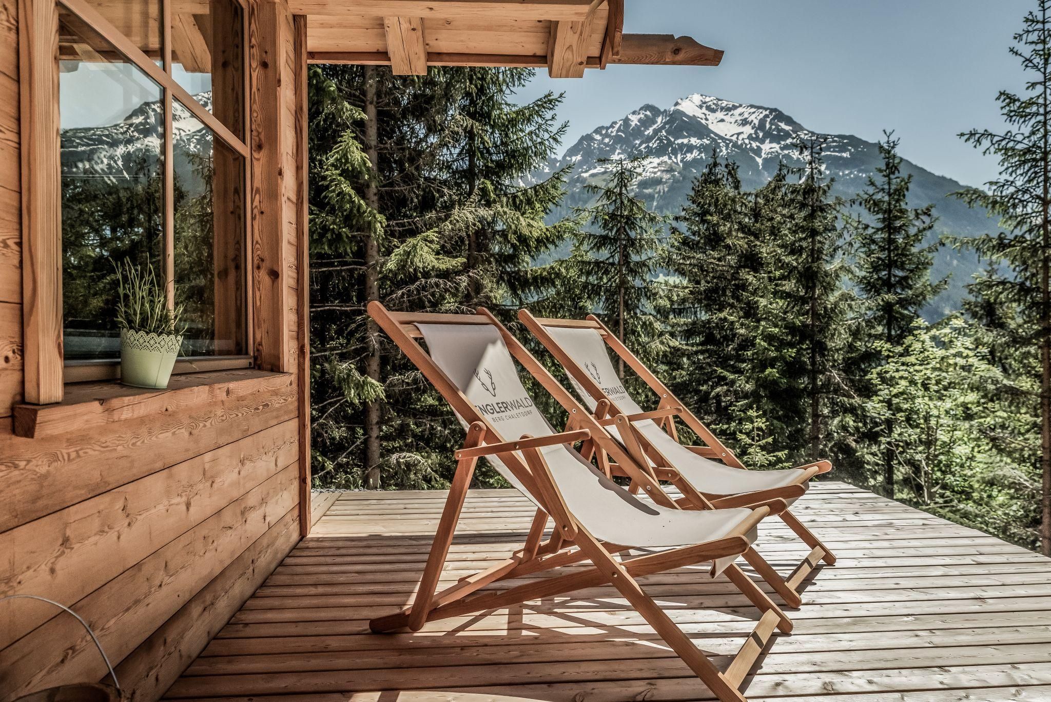 Eine gemütliche Terrasse mit zwei Liegestühlen blickt auf einen bewaldeten Berg. Im Hintergrund sind schneebedeckte Gipfel sichtbar.