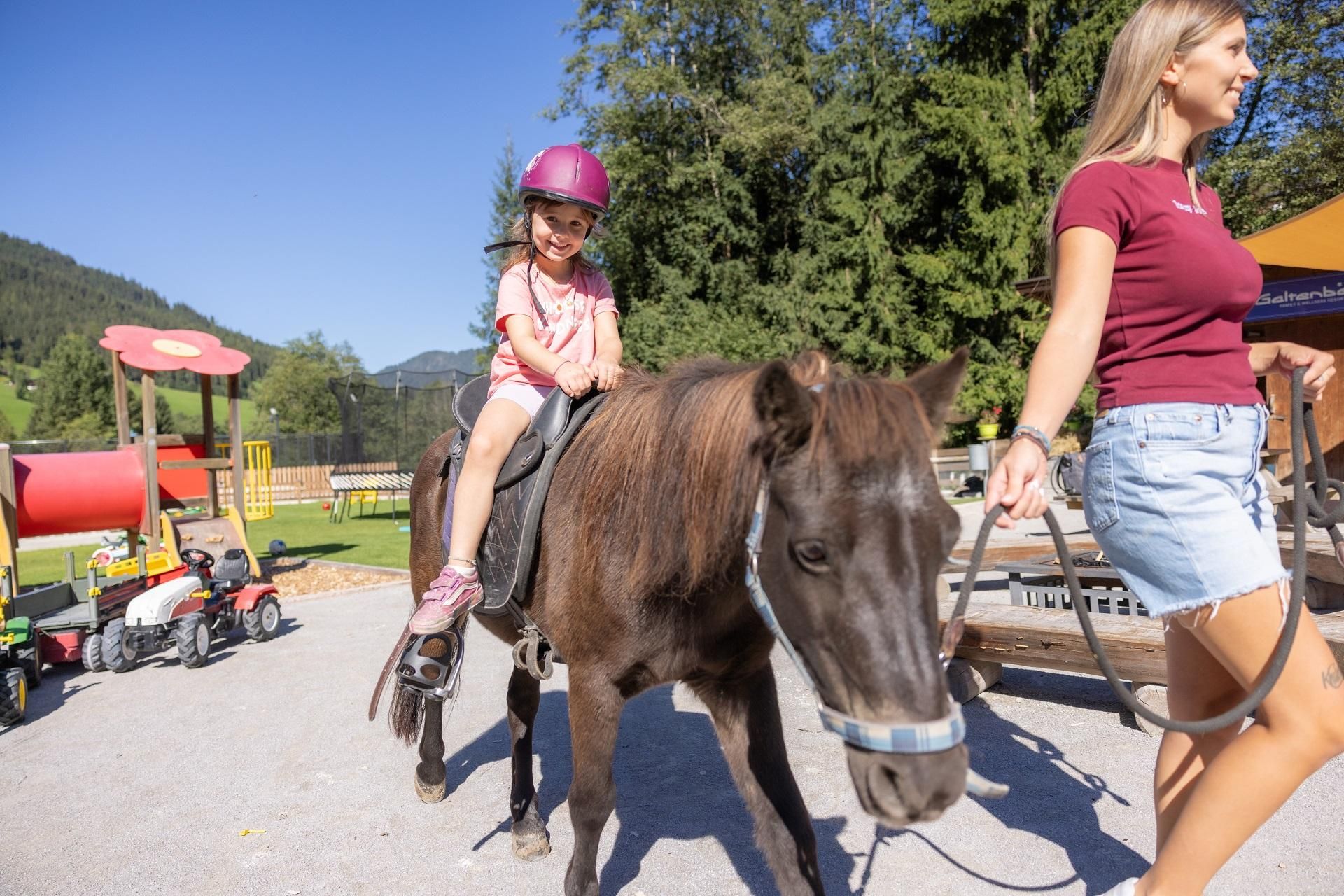 Ein kleines Mädchen reitet auf einem Pony und trägt einen Helm. Eine Frau führt das Pony auf einem Spielplatz mit grüner Umgebung.