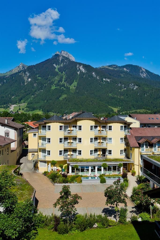 Ein sonniges Hotelgebäude umgeben von grünen Landschaften und Bergen. Der klare blaue Himmel ergänzt die frische Atmosphäre.