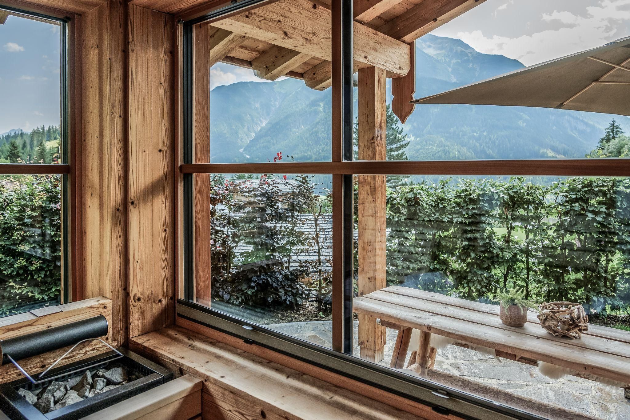 Ein gemütlicher Innenraum mit großen Fenstern, die einen Blick auf die Berge und grünes Terrain bieten. Ein rustikaler Tisch und moderne Elemente schaffen eine einladende Atmosphäre.