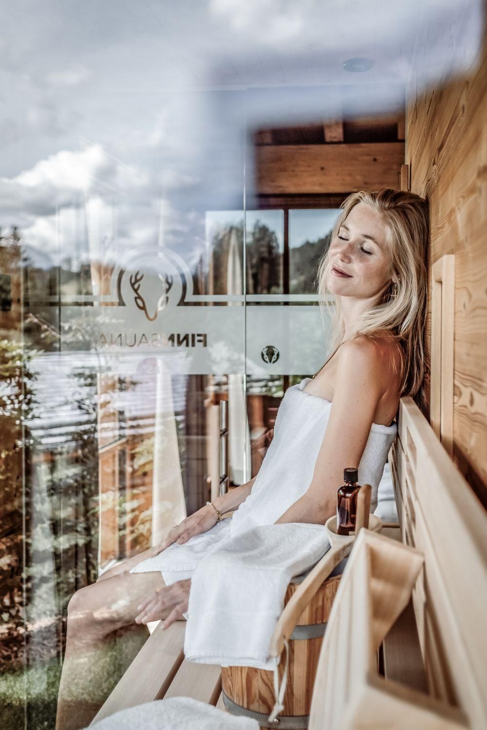 Eine entspannte Frau sitzt in einer Sauna und genießt die Ruhe. Durch das Fenster ist eine schöne Aussicht auf die Natur zu sehen.