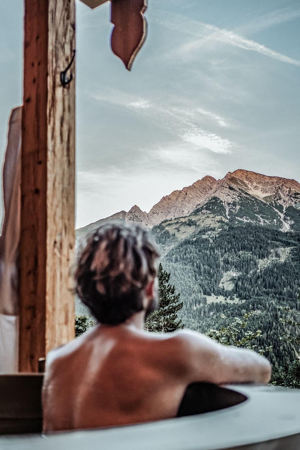 Eine Person sitzt entspannt in einem Hot Pot und blickt auf die majestätischen Berge. Der Himmel ist klar und die Natur wirkt friedlich.
