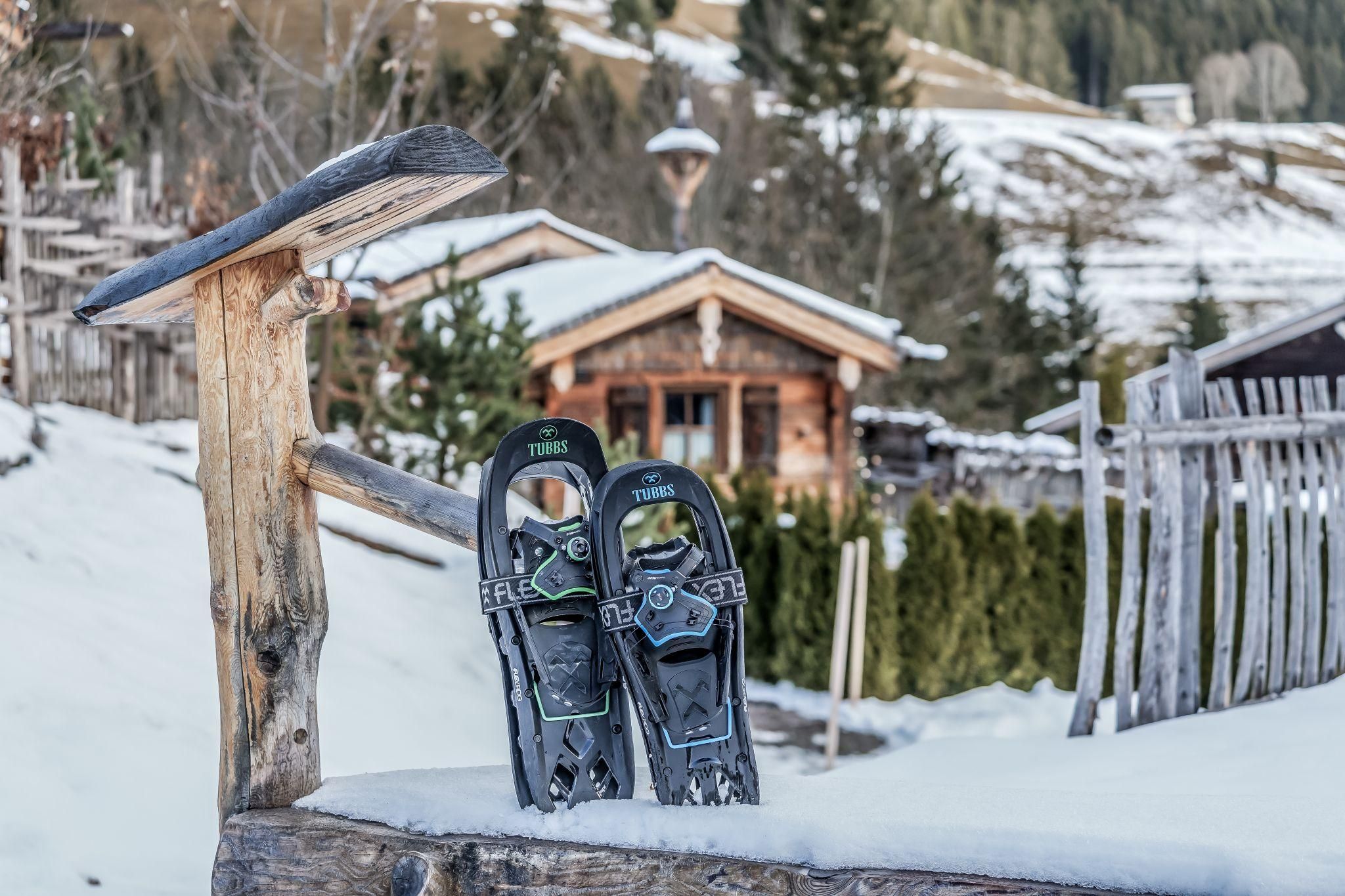 Zwei Schneeschuhe stehen auf einem Holzgeländer im Schnee. Im Hintergrund ist eine gemütliches Chalet und verschneite Bäume zu sehen.