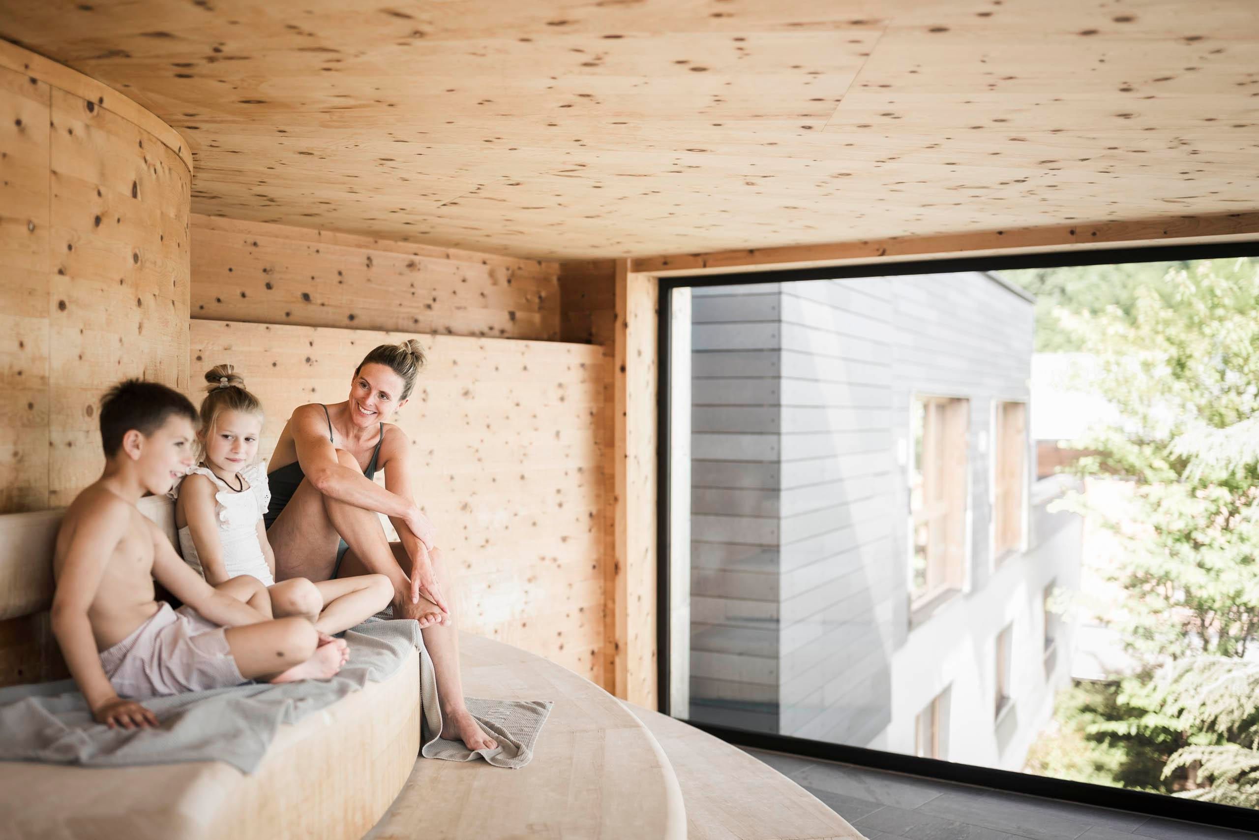 Eine Familie entspannt sich in einem modernen, hellen Raum mit Holzverkleidung. Sie sitzen auf einer komfortablen Sitzgelegenheit und genießen die Aussicht durch große Fenster.