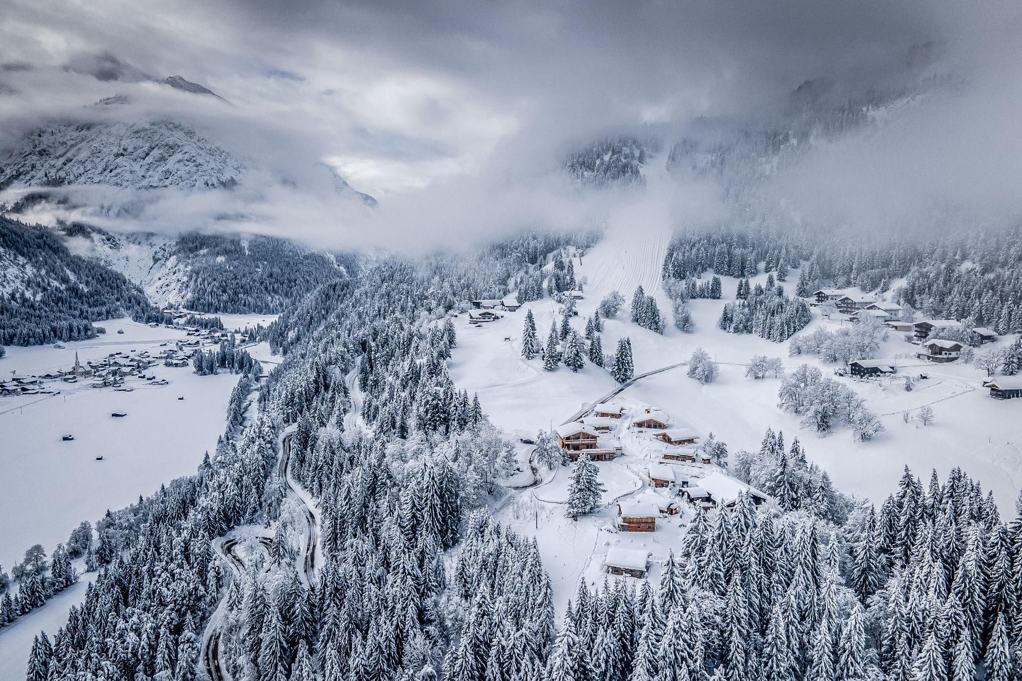 Eine schneebedeckte Landschaft mit Bergen und Wald. In der Ferne sind einige Chalet und ein zugefrorener See zu sehen.