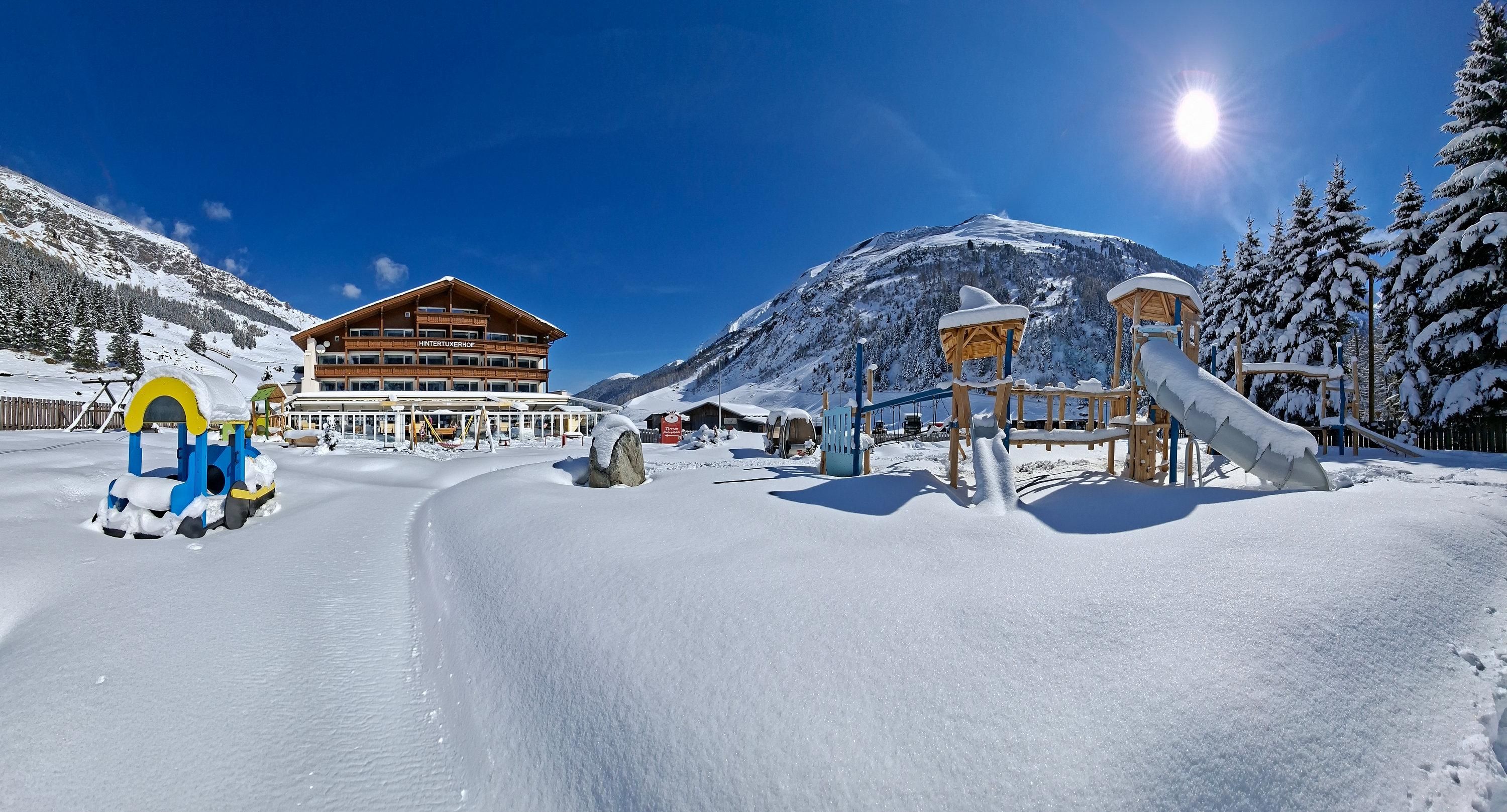 Eine verschneite Winterlandschaft mit einem Spielplatz und einem Gebäude im Hintergrund. Der Himmel ist klar und die Sonne strahlt.