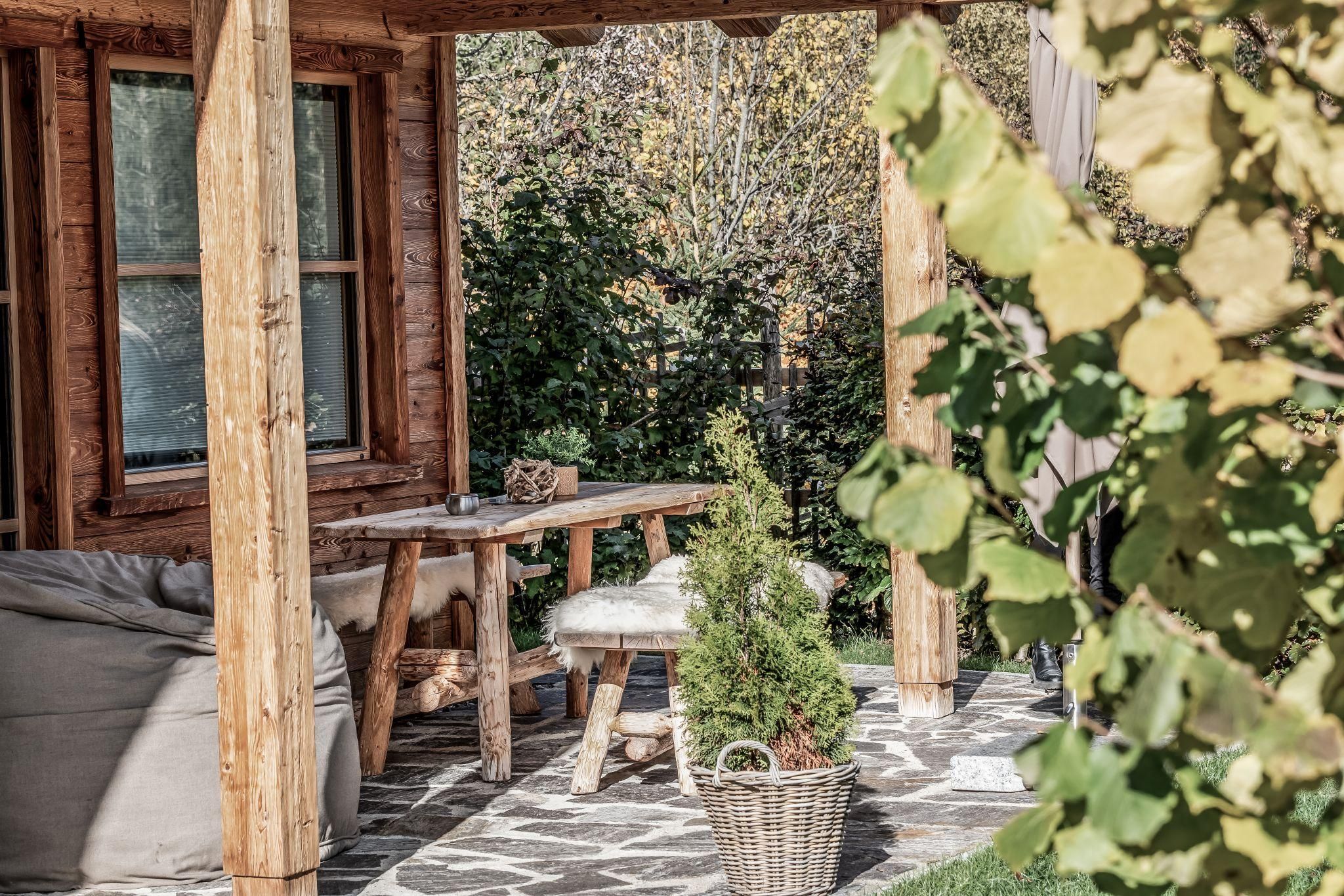 Eine gemütliche Terrassenansicht mit Holzsäulen und einem Tisch. Im Hintergrund sind Pflanzen und Bäume zu sehen.