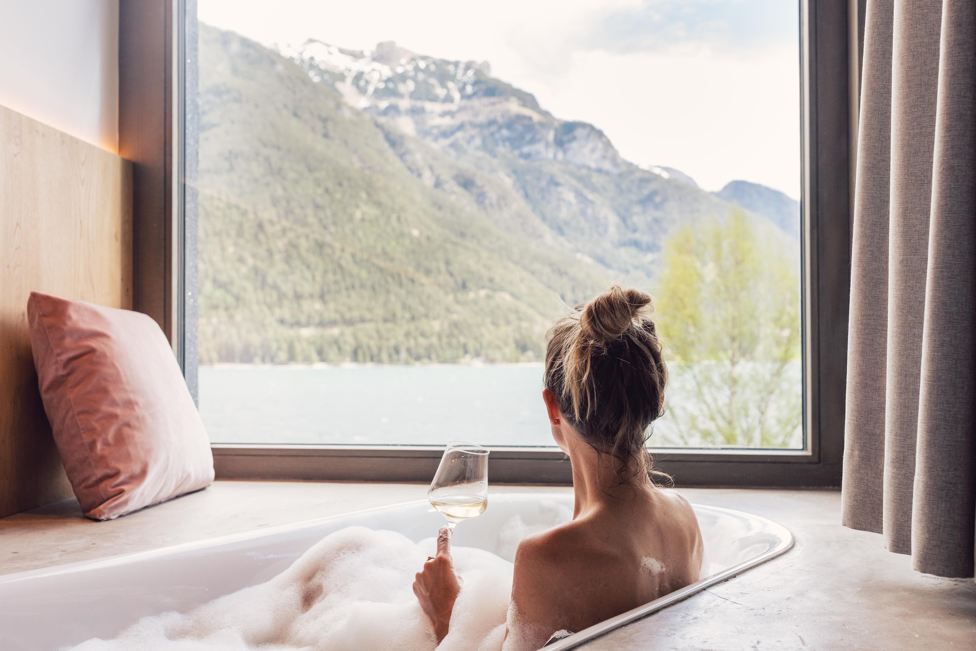 Eine Person sitzt in einer Badewanne und genießt einen Ausblick auf die Berge und einen See. Sie hält ein Glas in der Hand und umgeben von Schaum und Kissen.