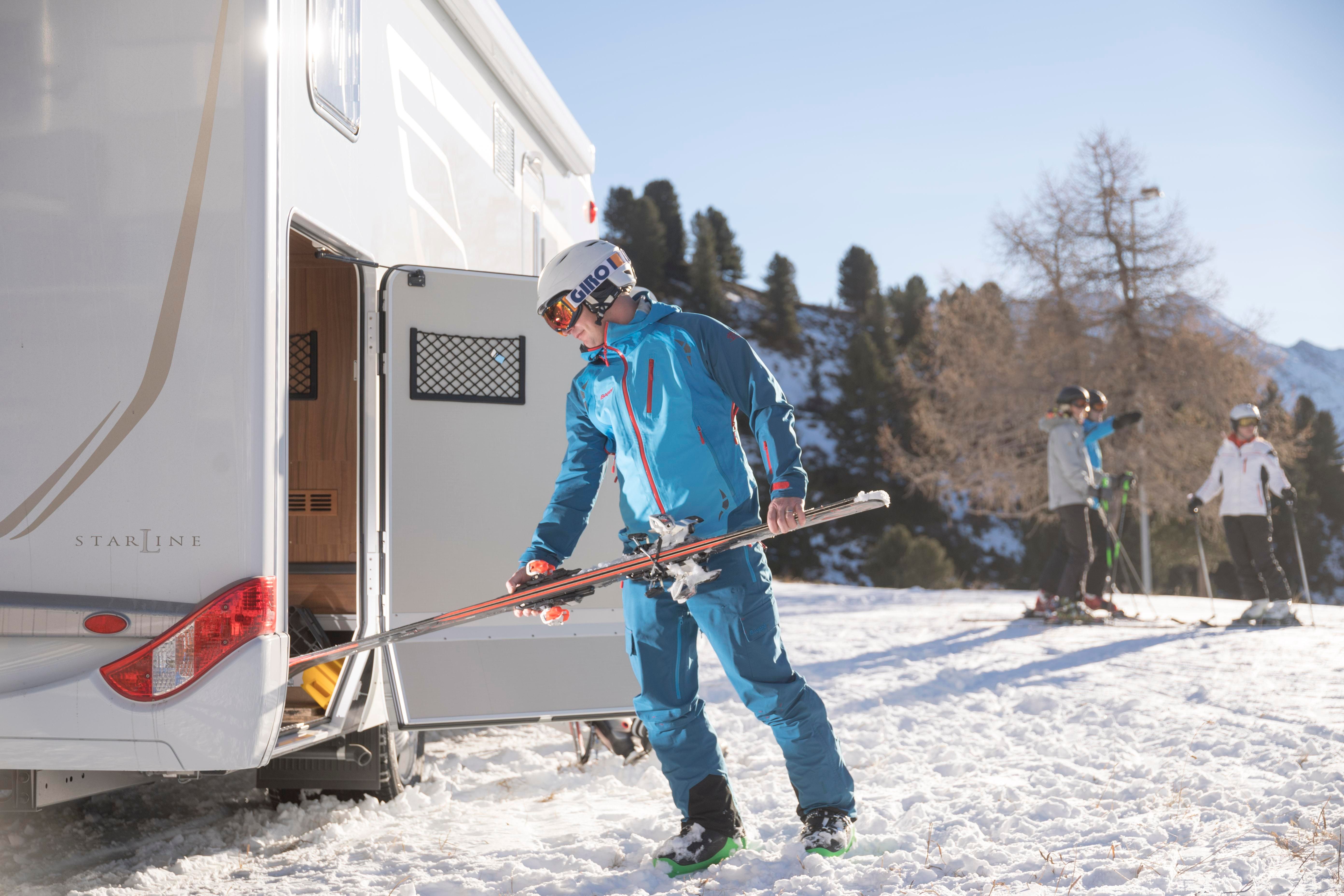 Ein Skifahrer in blauer Ausrüstung bereitet sich vor, seine Skier aus einem Wohnmobil zu holen. Im Hintergrund sind weitere Skifahrer auf einer schneebedeckten Piste zu sehen.