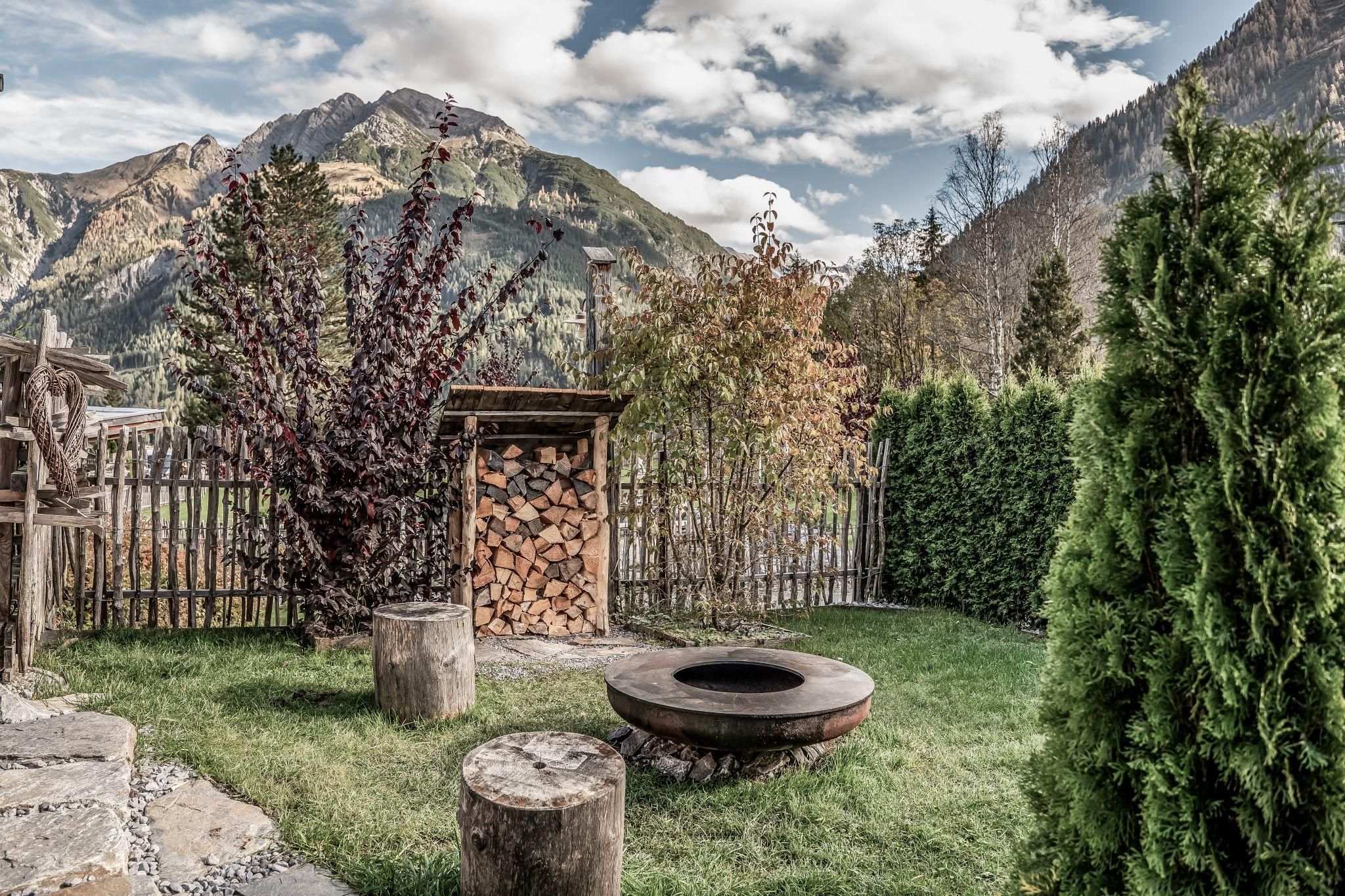 Ein idyllischer Garten mit eineGrillschale, umgeben von Bäumen und einem Holzstapel. Im Hintergrund erheben sich majestätische Berge unter einem bewölkten Himmel.