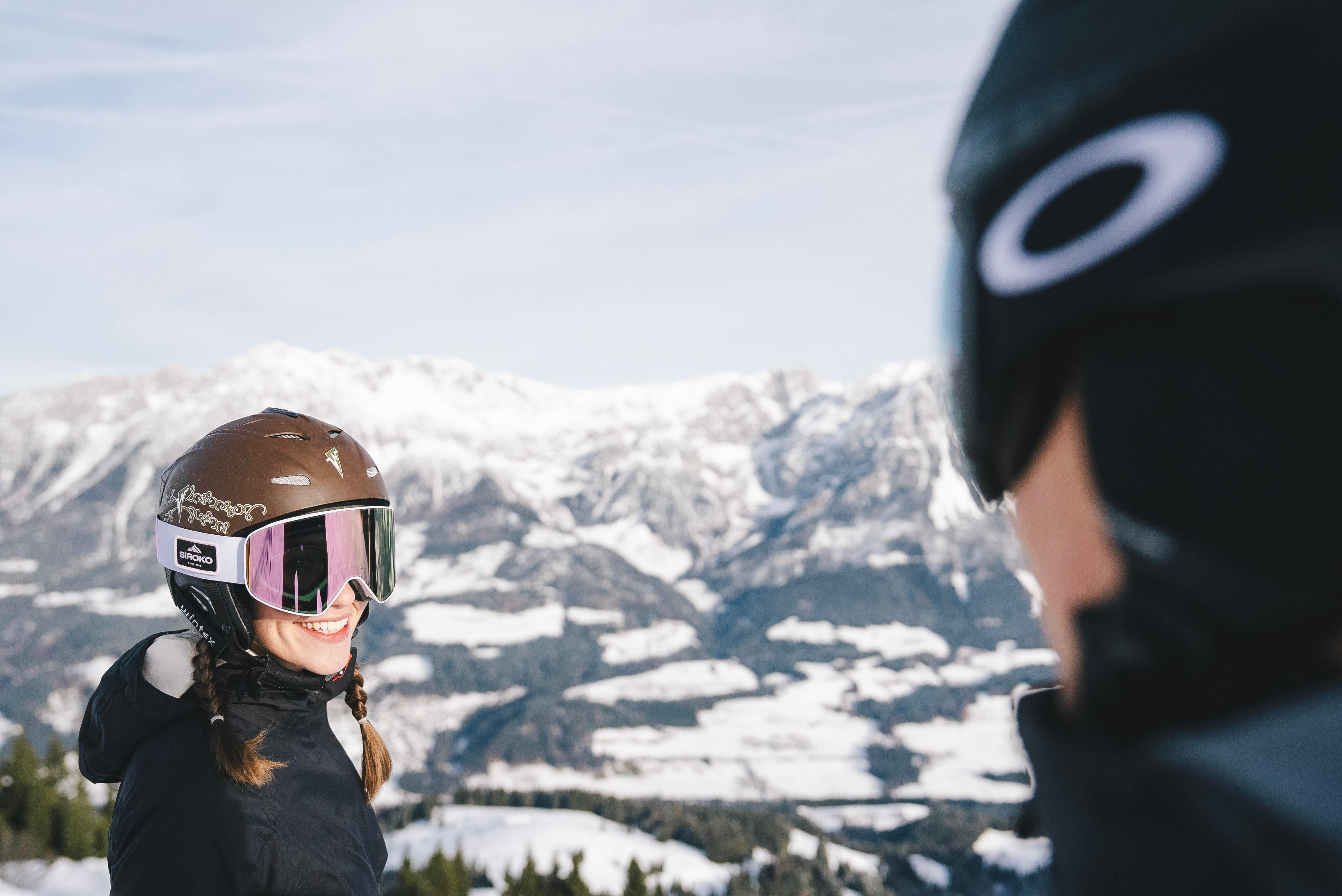 Zwei Skifahrer lächeln sich auf einem schneebedeckten Berg gegenüber. Im Hintergrund sind majestätische Berge und ein klarer Himmel zu sehen.