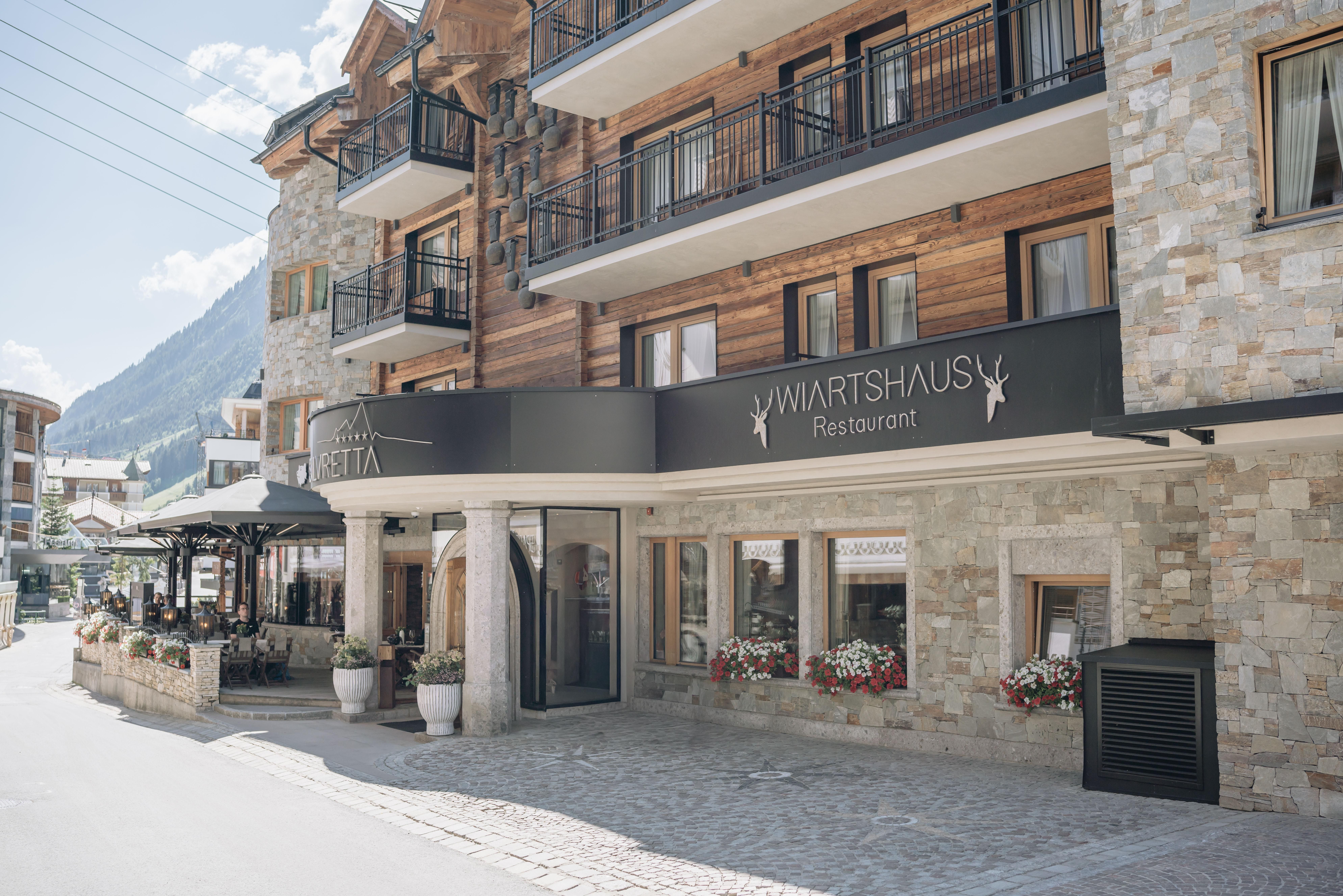 Ein modernes Restaurant mit einer einladenden Fassade aus Holz und Stein. Draußen stehen Tische mit Blumen und die Umgebung ist von Bergen und einem blauen Himmel umgeben.
