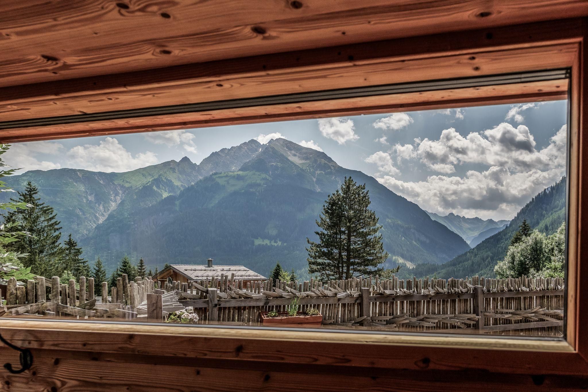 Eine beeindruckende Berglandschaft mit hohen Gipfeln und bewaldeten Hängen. Der Blick durch ein Fenster zeigt eine ruhige Atmosphäre mit einem kleinen Chalet im Vordergrund.