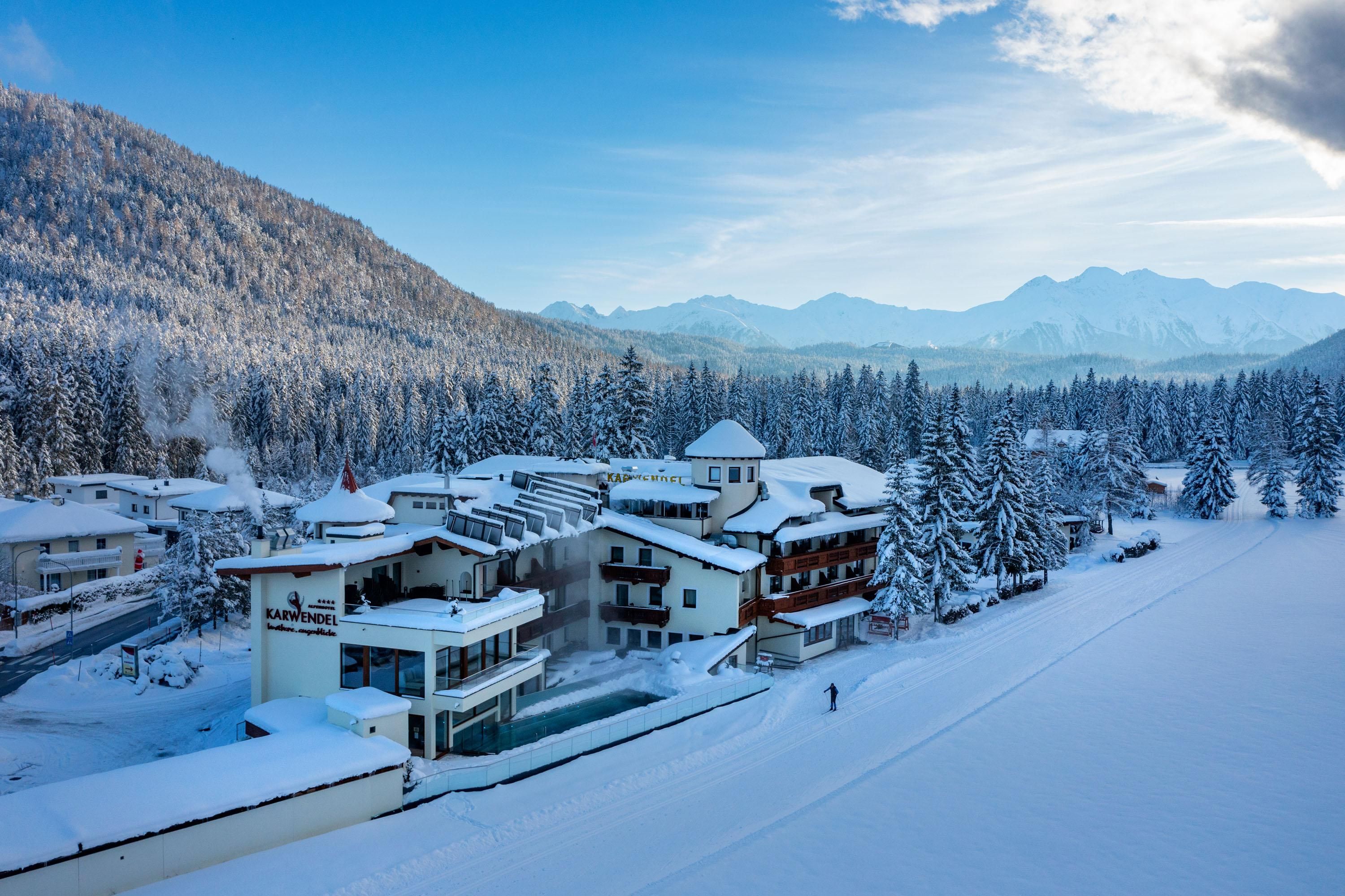 Eine idyllische winterliche Landschaft mit schneebedeckten Bäumen und Bergen im Hintergrund. Im Vordergrund sind gemütliche Gebäude zu sehen, die in der ruhigen Umgebung liegen.