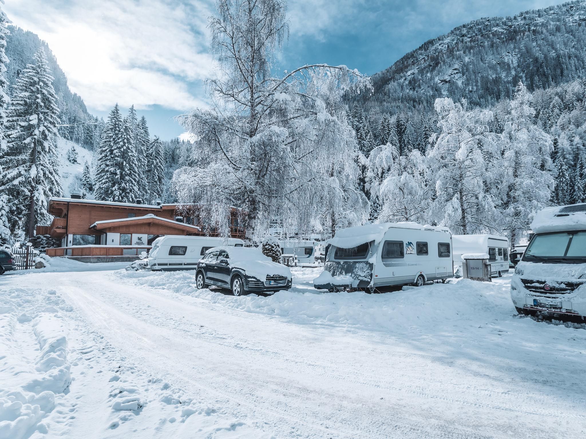 Ein verschneiter Campingplatz mit Wohnwagen und parkenden Autos. Im Hintergrund sind schneebedeckte Berge und Bäume zu sehen.