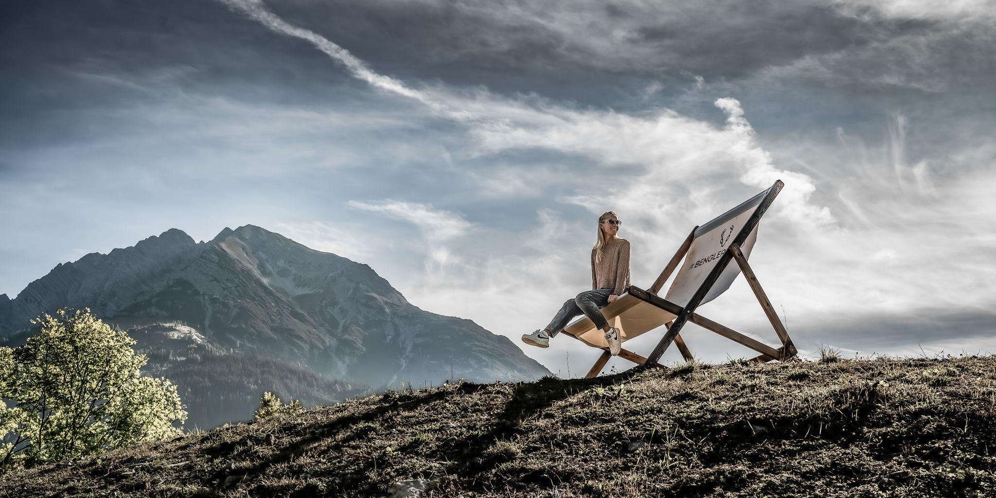 Eine Frau sitzt entspannt auf einer Liege in der Natur. Im Hintergrund sind Berge und ein bewölkter Himmel zu sehen.