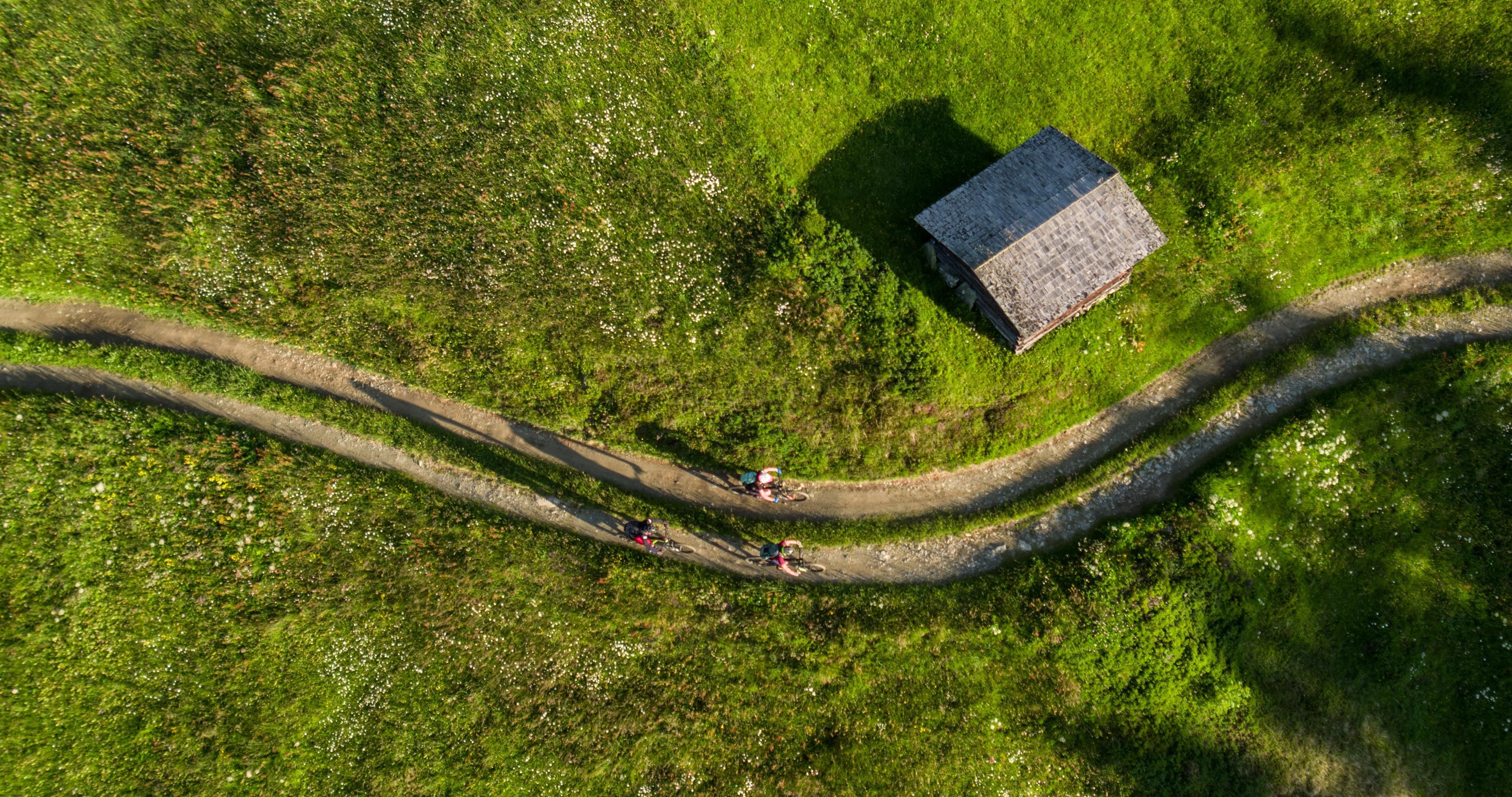 Eine malerische Landschaft mit grünen Wiesen und einem kleinen Holzhaus. Auf einem gebogenen Weg fahren Radfahrer vorbei.
