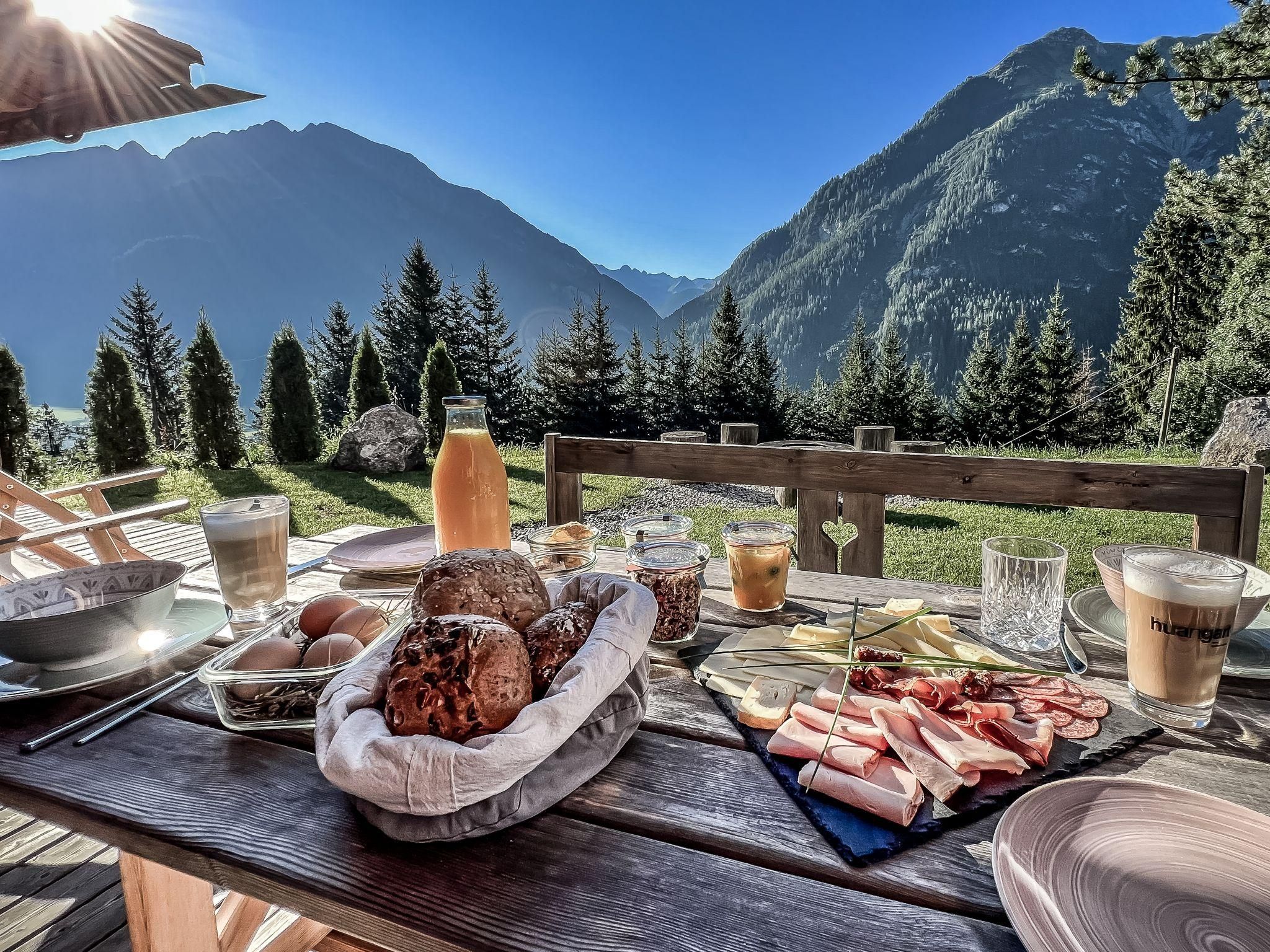 Ein schöner Frühstückstisch im Freien mit Bergen im Hintergrund. Das Tischgedeck umfasst Brötchen, Aufschnitt und Getränke.