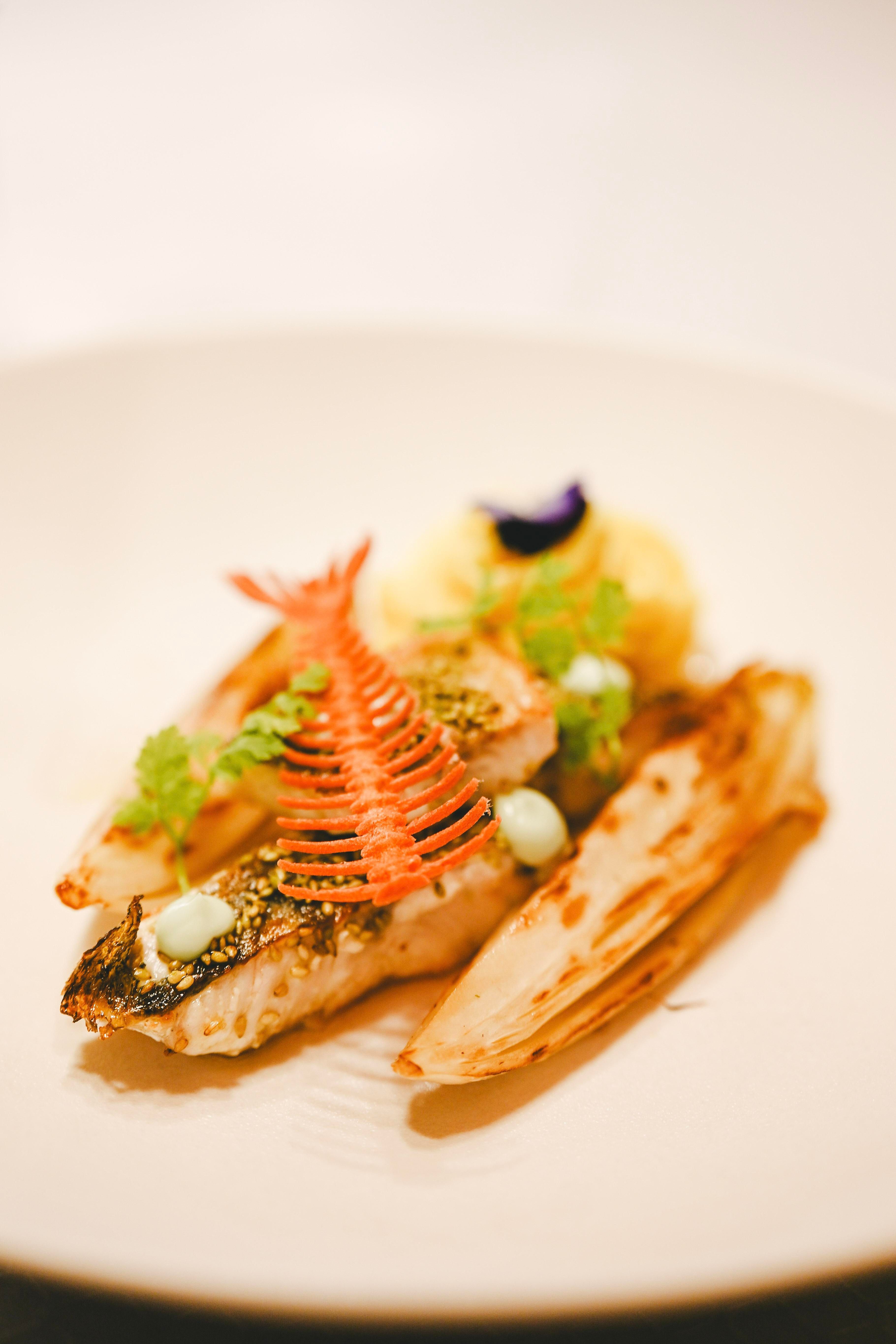 Ein kunstvoll angerichteter Fisch auf einem weißen Teller, garniert mit frischen Kräutern und bunten Dekorationen. Dazu sind gegrillte Gemüsebeilagen zu sehen.